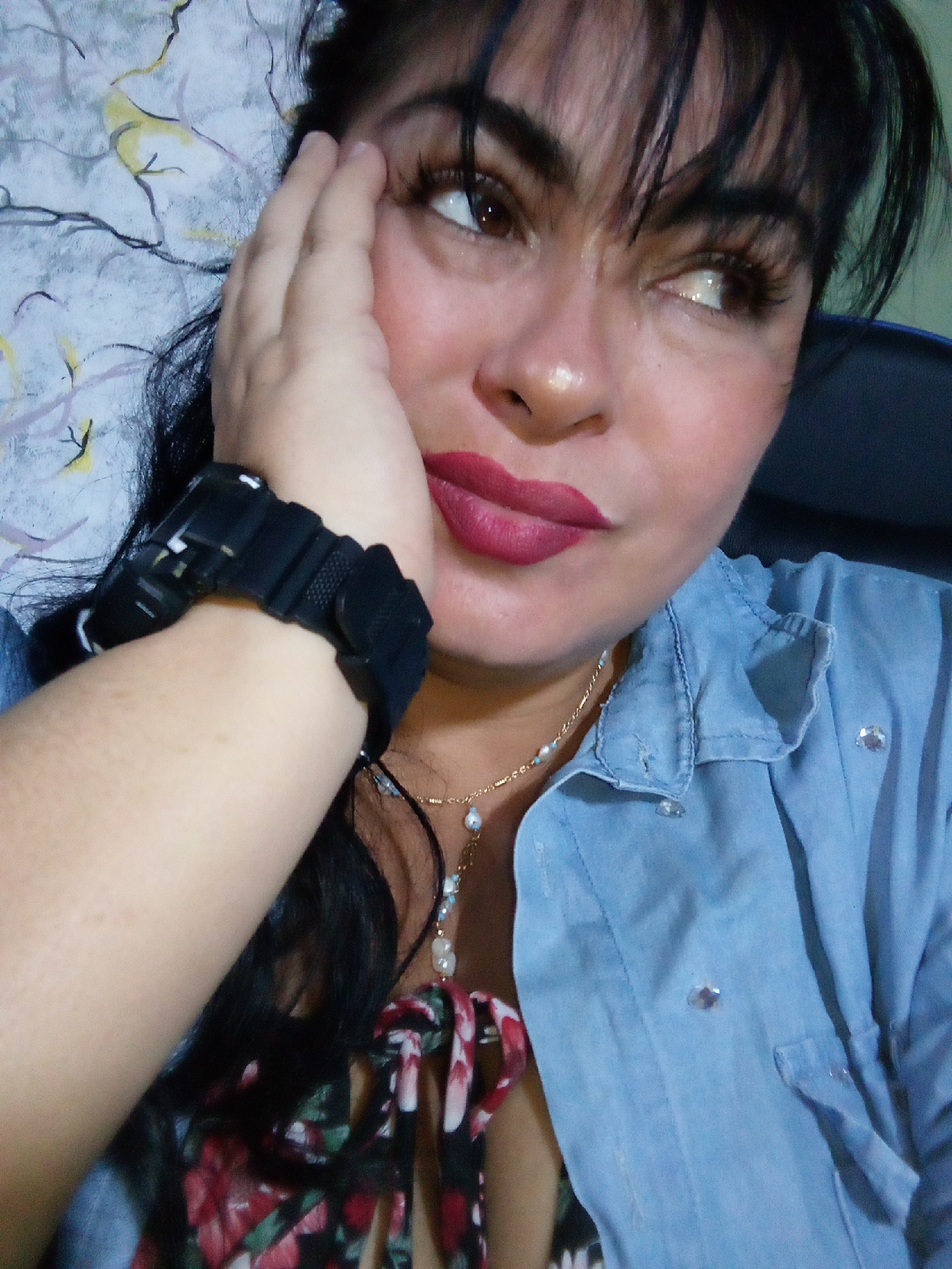This close-up portrait features a woman with dark hair and bangs, captured in what appears to be an indoor or shaded setting in Juan Griego, Venezuela. She gazes pensively towards her right (viewer's left), with her left hand gently resting against her cheek, suggesting a moment of thought or reflection.
She is dressed in a light blue denim-style shirt or jacket, adorned with small, sparkling crystal-like buttons. Beneath it, a colorful top with a floral pattern in red, white, and green on a dark background is visible. Her accessories include a delicate gold chain necklace with small white pearls and light blue beads, and a bulky black digital watch worn on her left wrist.
The woman's makeup is notable, featuring defined dark eyebrows, prominent eyelashes (likely false), and a striking reddish-pink lipstick. A subtle highlight brightens the bridge of her nose. The background behind her left side displays a light-colored fabric with an abstract, squiggly design in gray, white, yellow, and black hues, while a dark object, possibly a chair back, is partially visible behind her right shoulder. The soft, even lighting does not indicate a specific time of day. No visible text is present in the image.