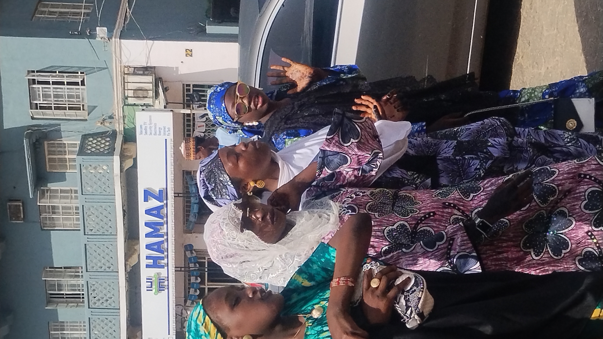 In Jos, Nigeria, four women are seen outdoors. They are dressed in colorful, patterned fabrics and head coverings. They are positioned in a way that they appear to be lying or sitting on top of each other. One of the women is wearing sunglasses and has intricate patterns on her hands. In the background is a building with the word "HAMAZ" prominently displayed and a number 22. The building appears to be a commercial or residential space. The scene is captured in daylight, suggesting an outdoor setting during daytime.