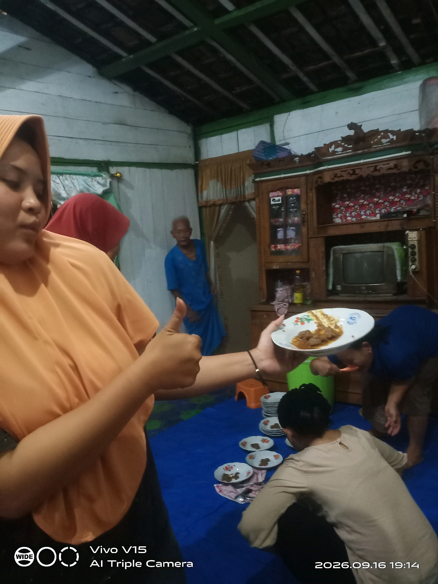 In a dimly lit room with white wooden walls and a dark, sloped ceiling, a group of people are gathered for a meal. In the foreground, a person wearing an orange hijab and shirt holds up a white plate with food, giving a thumbs-up. Behind them, another person in a red hijab is partially visible. To the right, a wooden cabinet with glass doors and intricate carvings stands against the wall, holding various items, including an old television set. Below the cabinet, a person in a blue shirt is seen bending down, seemingly eating or serving food. Several plates with food are arranged on a blue mat on the floor, where another person in a beige shirt is also seated. The atmosphere suggests a casual, indoor gathering. The bottom of the image displays a timestamp, "2026.09.16 19:14," and a "Vivo V15 AI Triple Camera" logo.