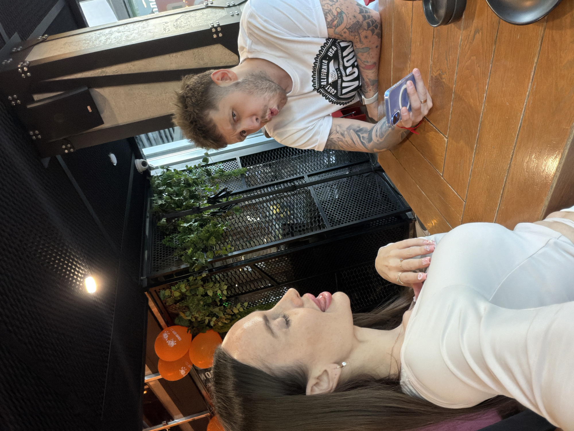 This detailed image captures two individuals, a woman and a man, seated at a light brown wooden table within a Tokio City restaurant in Saint Petersburg, Russia.
In the foreground on the left, a woman with long, straight dark brown hair is depicted in profile, facing right. She has fair skin with visible freckles on her forehead and cheekbones. Her eyes are closed, and her tongue is extended out of her mouth in a playful manner. She is wearing a light-colored, possibly white or off-white, long-sleeved top with a subtle v-neck. A delicate necklace is visible, along with a small stud earring in her left ear. Her right hand, adorned with multiple rings and nails featuring intricate designs, rests near her chest.
In the midground on the right, a man with short, textured light brown hair and a beard/stubble is seated, facing the camera. He is making a puckering or "fish-face" expression with his lips. He wears a white short-sleeved t-shirt with a prominent black circular graphic on the chest that reads "COAST CALIFORNIA USA RACING ORANGE 1991 VCON". Both of his arms are extensively covered in various tattoos. A white smartwatch is visible on his left wrist, and his right hand, resting on the table, holds a purple-cased smartphone.
The immediate environment includes the polished wooden table. On the table to the right are two stacked dark, round dishes, with another similar dish partially visible further right. The background of the restaurant features a dark, possibly black, textured ceiling with a single bright overhead light fixture that casts a patterned reflection. Below the ceiling, a black mesh-like partition is adorned with artificial green foliage. Several orange balloons are visible in the upper left background, one of which clearly displays "TOKIO-CITY". A large, multi-faceted dark pillar with metallic decorative elements stands behind the man, with a black speaker mounted on it. A small, white dome-shaped security camera is mounted on a structure in the upper left. Large windows or glass partitions are visible in the far background, suggesting an indoor setting with external light. Both individuals exhibit playful facial expressions, indicating a lighthearted interaction.