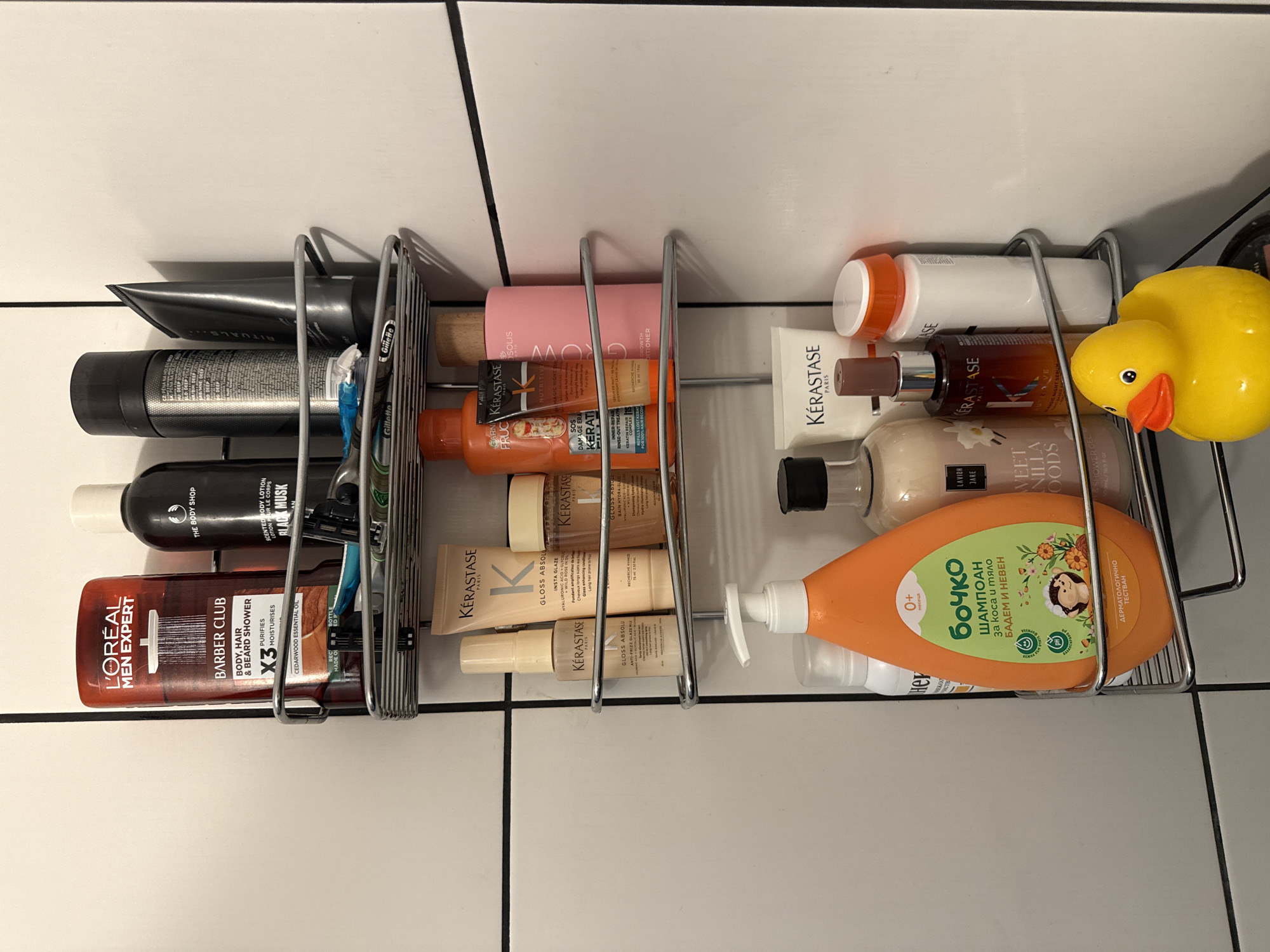 The image shows a close-up, eye-level view of a multi-tiered shower caddy filled with various toiletries and personal care products. The shower caddy is mounted on a white tiled wall, with black grout lines forming a grid pattern. The caddy itself is made of chrome-plated wire.
On the top shelf, from left to right, are a large red bottle of L'Oréal Men Expert Barber Club Body, Hair & Beard Shower Gel, followed by a black bottle of The Body Shop Scented Body Lotion in Black Musk. Next to it is a black tube of what appears to be a face mask or scrub, and a smaller black tube with a silver cap. A blue and silver razor and a few small tubes are also visible on this shelf.
The second shelf from the top contains several Kérastase products. These include small bottles, tubes, and a larger pink bottle of Kérastase Nutritive Bain Hydra-Gloss Conditioner.
The third shelf holds a large orange bottle of "Бочко" (Bochko) brand shampoo for babies, with a cartoon hedgehog on the label. Next to it is a clear bottle with a white pump top, and a dark bottle with a white label that reads "LAVISH JARE SWEET VANILLA WOODS SHOWER GEL". To the right of these are more Kérastase products and a white bottle with an orange cap. A yellow rubber duck is positioned on the lower part of this shelf, partially obscuring some of the products.
The image is well-lit, with no shadows obscuring the details of the items. The environment appears to be a bathroom. There is no information in the image to suggest its location in Plovdiv, Bulgaria.