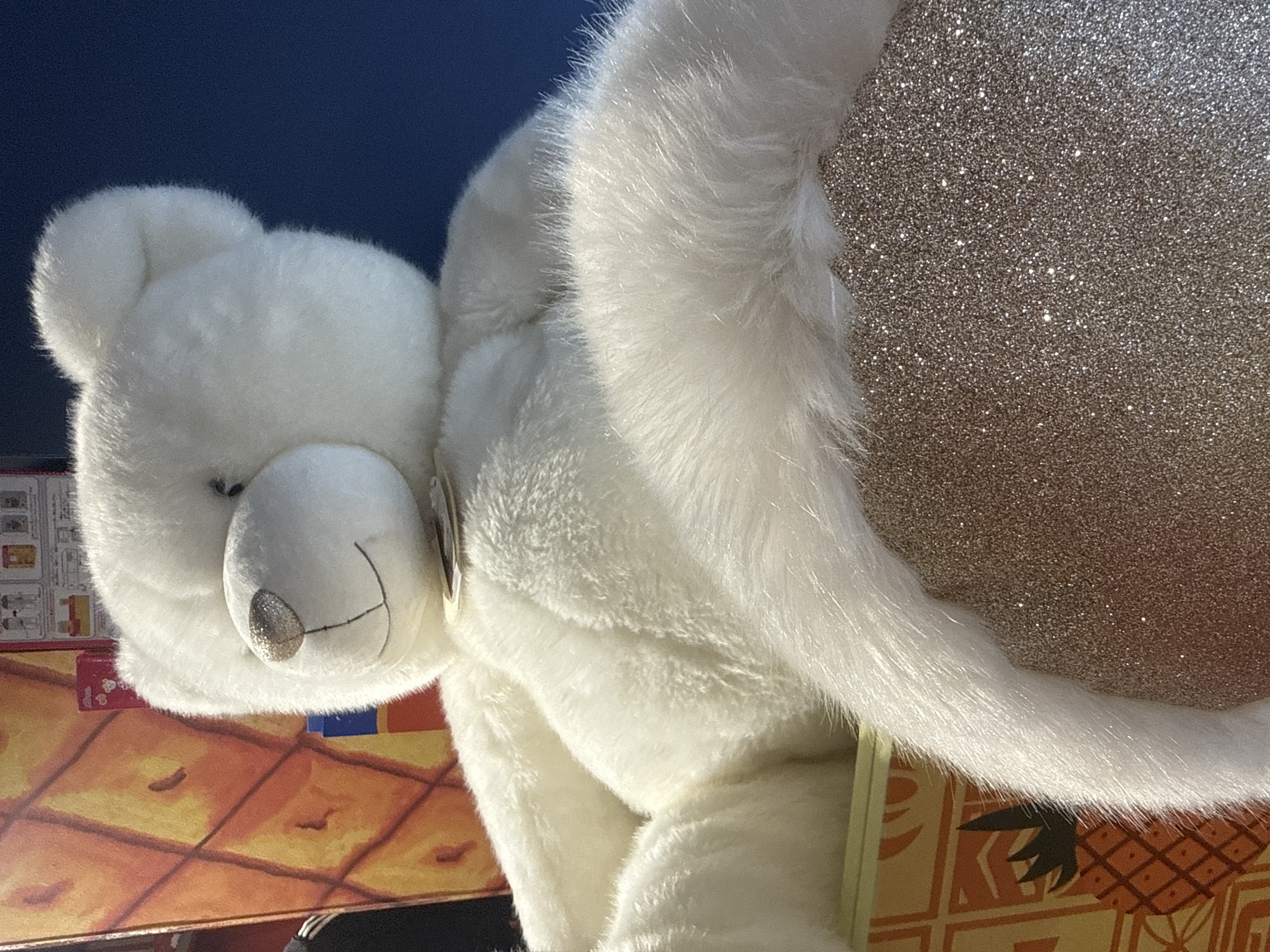 A close-up, eye-level shot depicts a white teddy bear holding a glittery ball. The teddy bear has white fur and a silver nose. The glittery ball is silver-toned with a dense, shimmering texture. The background includes a patterned surface with orange and yellow hues, and a dark blue section. There are no visible indicators of the location as Yekaterinburg, Russia.