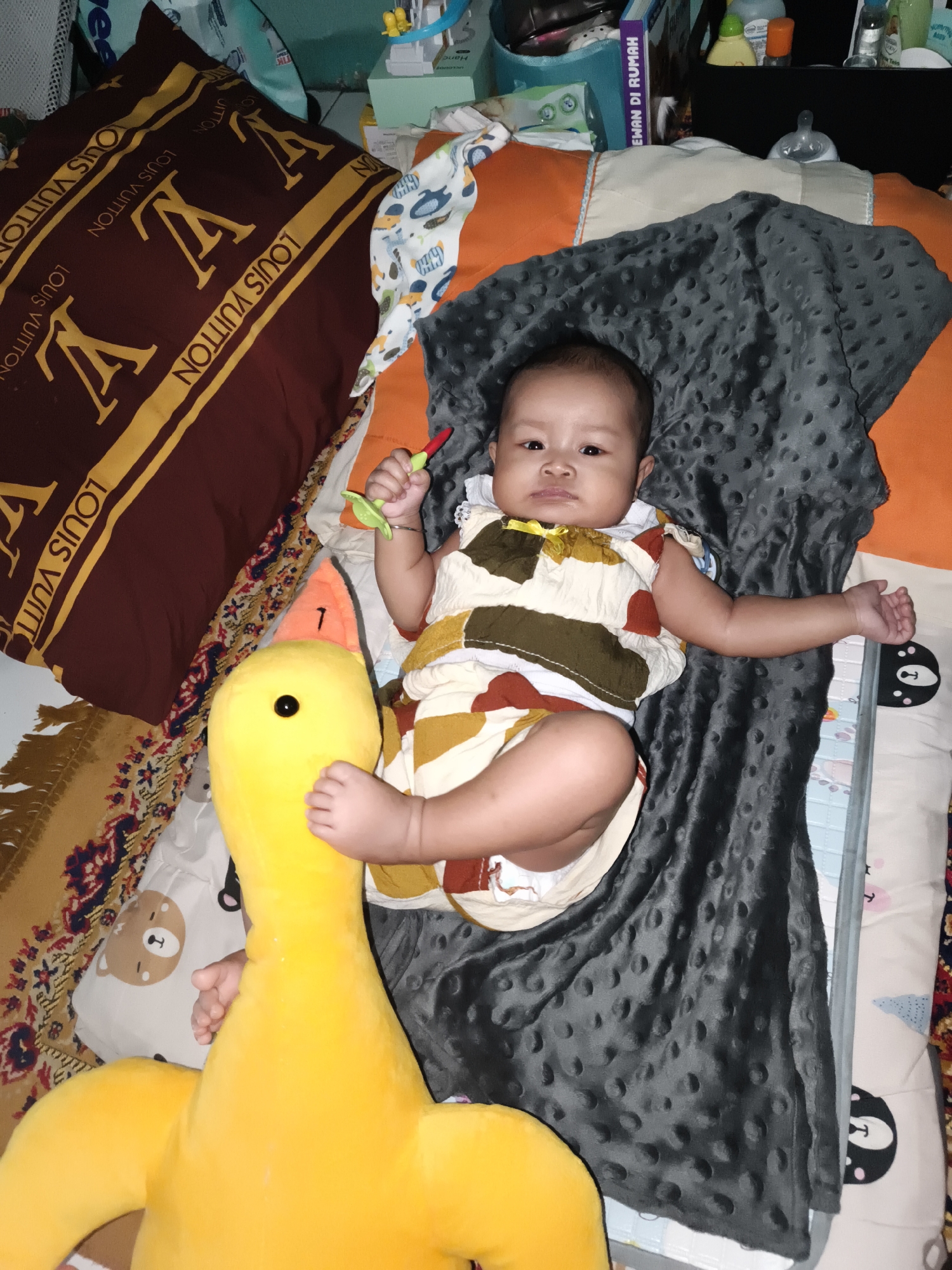 An infant lies on their back on a patterned bed in Barengkrajan, Indonesia. The infant is wearing a patterned onesie. To the left of the infant is a large, yellow stuffed duck. To the right of the infant is a gray, textured blanket. Behind the infant, there is a maroon pillow with a Louis Vuitton pattern, as well as other bedding and various items. The scene appears to be indoors, likely in a home.