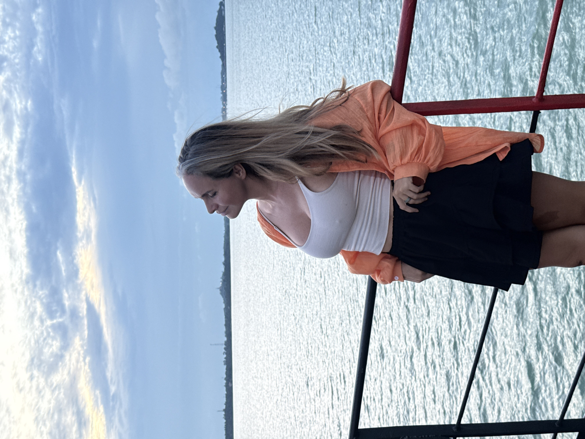 A woman with blonde hair, wearing an orange shirt over a white tank top and a black skirt, stands on a pier with her eyes closed, looking up at the sky. She is leaning on a red railing. The sea is in the background with a cloudy sky.