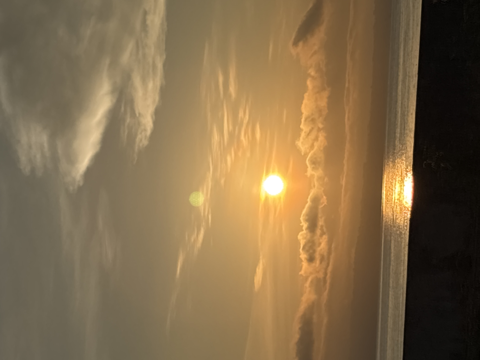 The image captures a serene sunset over the Davao Gulf, likely taken from a coastal area within Davao City, Philippines. The sun, a bright orb, is positioned in the middle of the frame, casting a warm, golden glow across the sky and the water. Wispy clouds are scattered throughout the sky, some catching the sunlight and appearing in shades of orange and yellow. The water of the gulf reflects the sun's light, creating a shimmering pathway of gold across its surface. In the foreground, a dark, silhouetted strip suggests the edge of a beach or a seawall, grounding the scene. The distant horizon shows a hazy outline of land, likely the coastline of Davao City or surrounding islands. The overall atmosphere is peaceful and tranquil, characteristic of a sunset over the ocean.