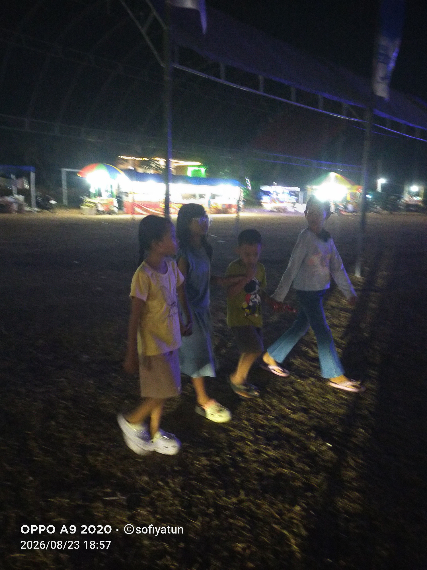 Four children are walking from left to right across a dark, outdoor area at night. The children are of varying heights and appear to be in motion, indicated by the blur in the image. The child on the far left is wearing a yellow shirt and a light-colored skirt or shorts, with white platform shoes. The second child from the left is wearing a blue dress and dark shoes. The third child is wearing a green shirt with a character print and dark shorts, with flip-flops. The child on the far right is wearing a light-colored long-sleeved shirt and blue pants, with pink flip-flops. In the background, there are illuminated stalls or tents, suggesting a market or event. The ground is covered in dark, possibly grassy or leafy material. The date and time stamp at the bottom left of the image indicates "2026/08/23 18:57". The location is identified as "OPPO A9 2020 - ©sofiyatun". Based on the information, the scene is likely taking place in the city of Cluwak, Indonesia.