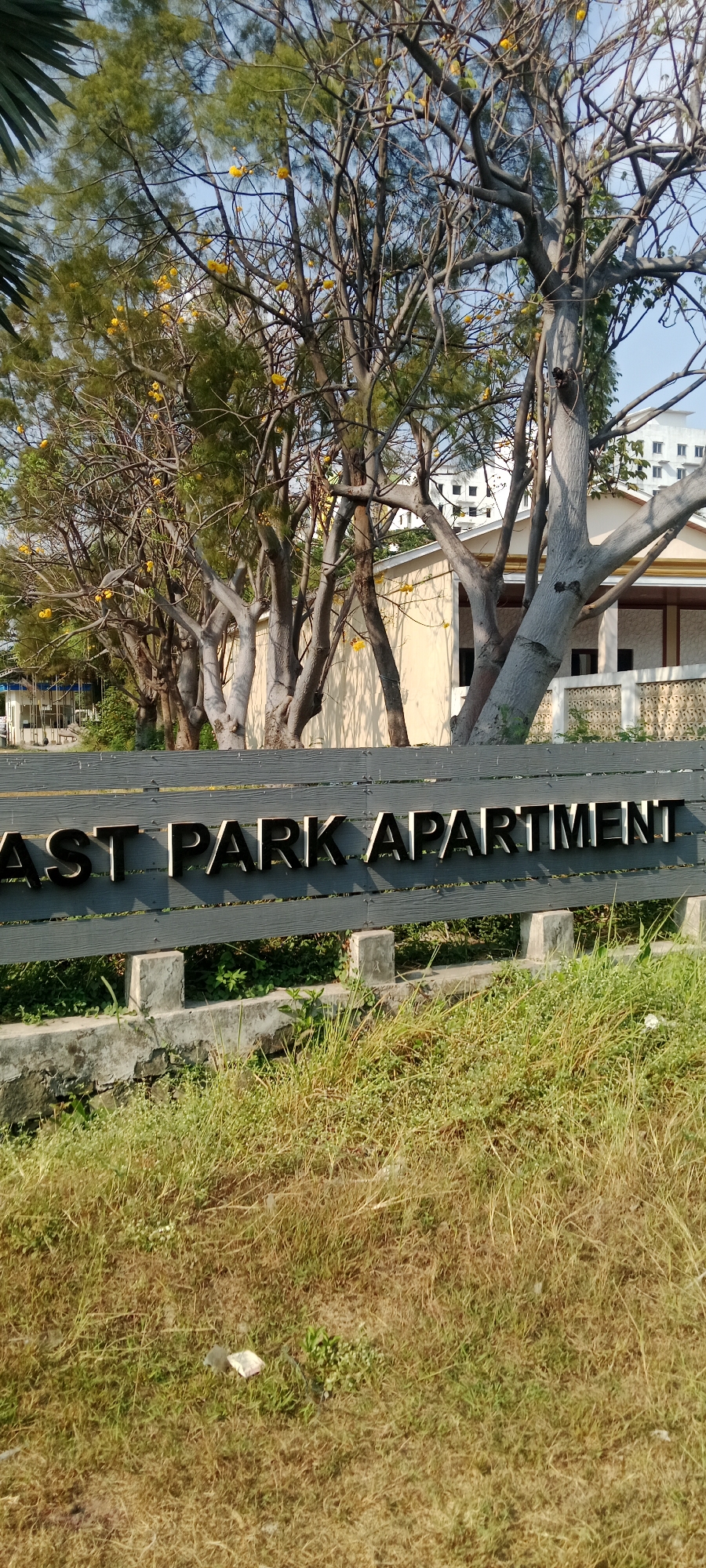 A weathered, dark gray wooden fence with weathered concrete posts stands in the foreground, displaying three-dimensional metallic letters spelling out "EAST PARK APARTMENT." The letters are a dark, reflective material that appears to be metal. Behind the fence, dry, yellowing grass and some green weeds grow. In the middle ground, several trees with thin branches and sparse green foliage are visible, some bearing small yellow flowers. Behind the trees, a light-colored building with a brown roof and dark windows is partially obscured. In the far background, a white multi-story building with visible windows stands against a clear blue sky. The overall scene suggests an outdoor environment in a developed area, possibly a residential or commercial complex. There are no people visible in the image.
