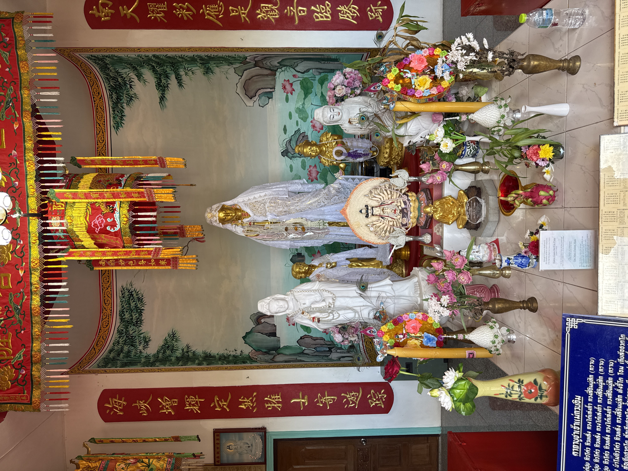 The image shows the interior of a shrine in Koh Phangan, Thailand. The shrine is decorated with red and gold banners, ornate lanterns, and floral arrangements. In the center, there are several statues of deities, including a large golden statue draped in white fabric and a white marble statue of a seated figure. Offerings of flowers, candles, and fruit are arranged in front of the statues. A painting of a landscape with bamboo and mountains adorns the back wall. The floor is tiled, and a blue informational sign is visible in the foreground. The overall atmosphere is one of reverence and spiritual significance.