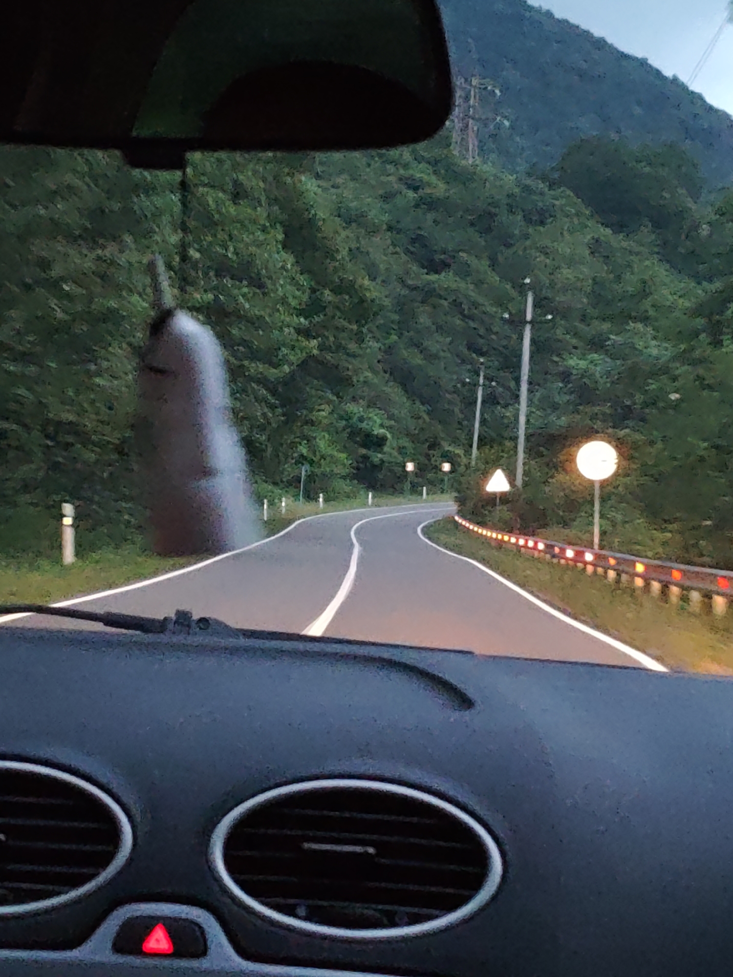 The image depicts a view from inside a vehicle, looking out at a winding road through a forest. The dashboard of the car is visible in the foreground, featuring two circular air vents and a red hazard light button illuminated. The road curves to the right, with white lines marking the lanes. On the left side of the road, a tall, dark, blurry object resembling a sculpture or natural formation stands against a backdrop of dense green trees. Further along the road, a guardrail with red reflective markers lines the right side. Several traffic signs are visible: a triangular warning sign and a circular sign with a bright light. The environment suggests a mountainous or hilly region with lush vegetation, likely in the late afternoon or early evening due to the subdued lighting. No people are directly visible in the scene, and no specific city or location is identifiable.