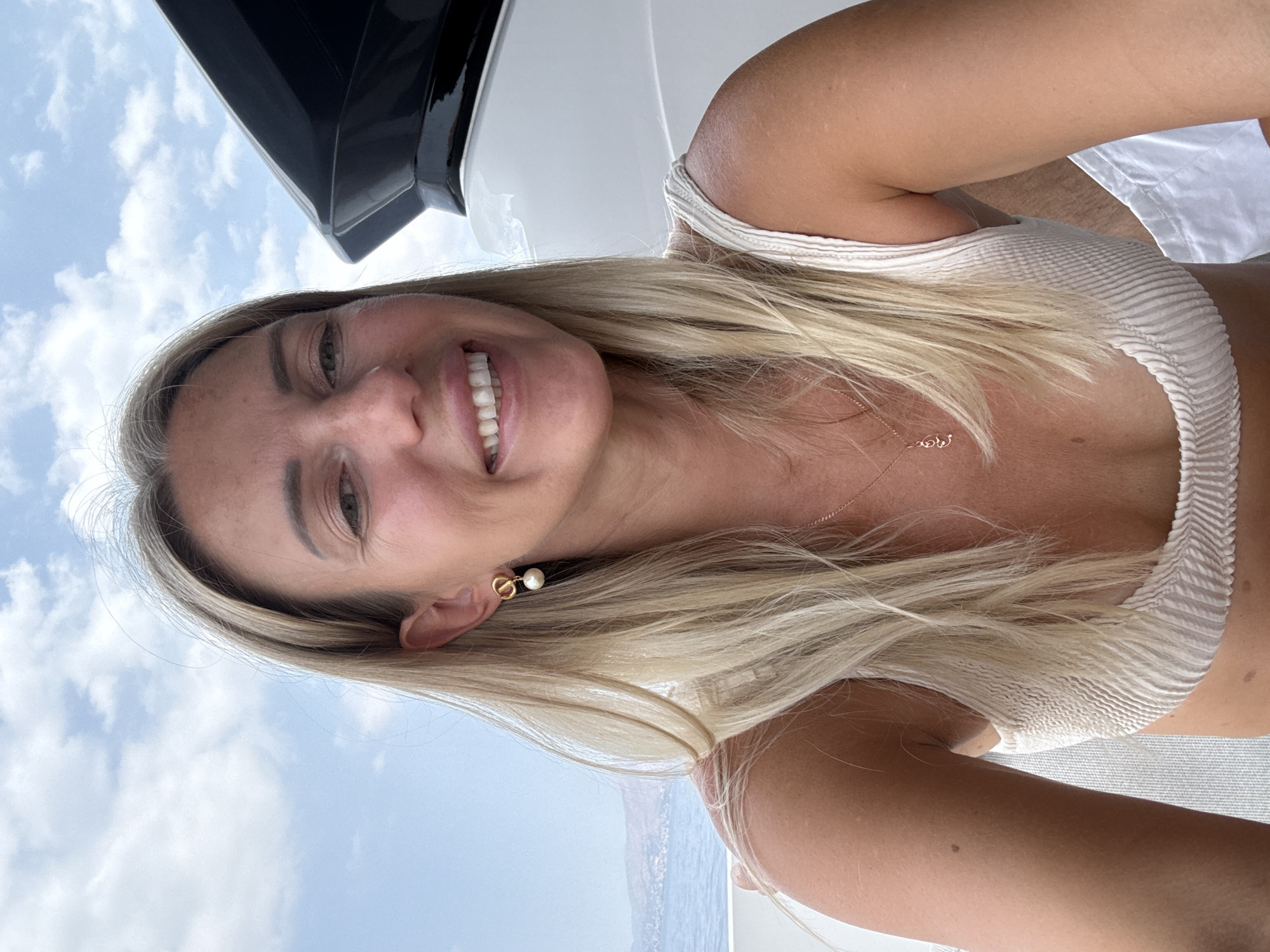 A woman with blonde hair smiles at the camera while on a boat. She is wearing a white bikini top and a necklace with a small pendant. The woman is outdoors, with a cloudy sky and the sea visible in the background. This image was taken in Agia Marina, Greece.