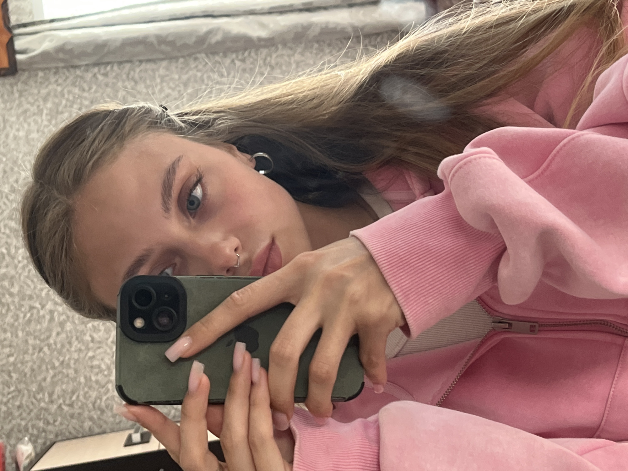 A young woman with blue eyes and light brown hair takes a selfie in what appears to be an indoor environment. She is wearing a pink hoodie and has a piercing in her nose. Her phone has a green case. The background is a patterned wall. There is no indication that this photo was taken in the city of Фоки, Russia.