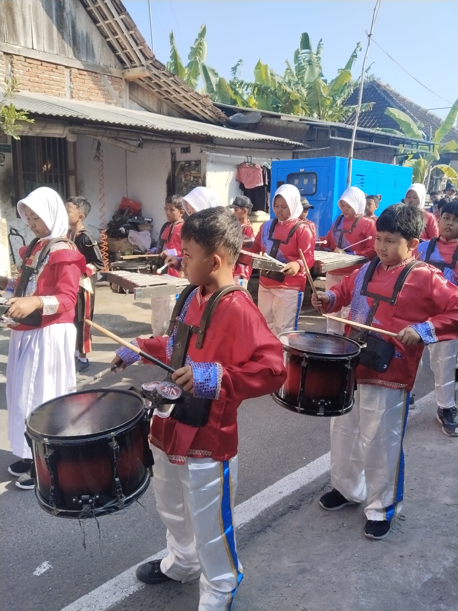 In a street scene in Gondang, Indonesia, a group of children in matching red and white marching band uniforms are participating in a parade. The children are carrying and playing various musical instruments. In the foreground, two boys are holding drums, with drumsticks in hand, suggesting they are actively playing. One boy, on the right, is wearing a red uniform with blue trim and white pants, his gaze directed forward. To his left, another boy in a similar uniform is also poised to play his drum.
Behind them, several other children are visible, some wearing white headscarves, also in uniform and holding instruments like xylophones. The background reveals a residential area with traditional Indonesian houses, some with exposed brick and wooden roofs, and lush green banana trees. A large, bright blue generator unit is also prominent in the background, suggesting it might be part of the parade's setup or a nearby facility. The overall atmosphere appears to be a daytime event under clear skies. The street is paved and has white lane markings.