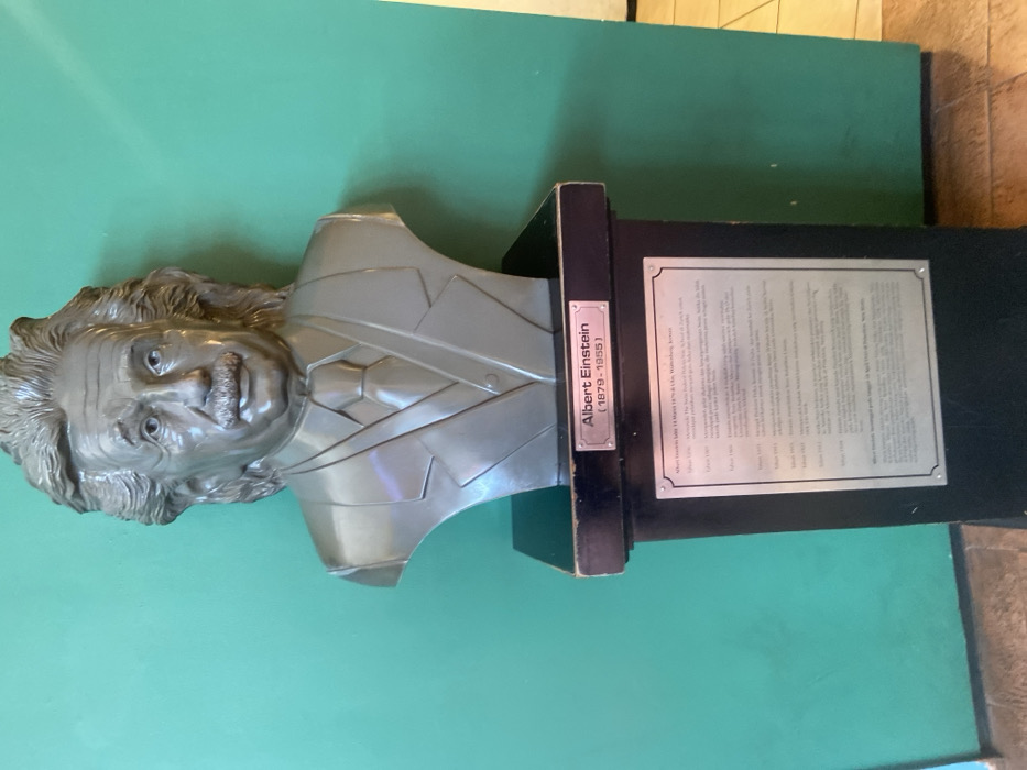 A bust of Albert Einstein, made of gray painted plaster, sits on a black pedestal. The bust depicts Einstein with a realistic likeness, wearing a suit with a tie. It is positioned against a green wall. The bust is located in a building in Padalarang, Indonesia, with coordinates of -6.852 latitude and 107.494 longitude. A black plaque on the pedestal states "Albert Einstein (1879-1955)" and a smaller plaque below lists key dates in his life. The pedestal is in a room with tiled flooring, with a green wall on one side and a doorway visible on the right side. There are no people visible in the image. The room appears to be a public space, possibly an educational institution or a museum.
