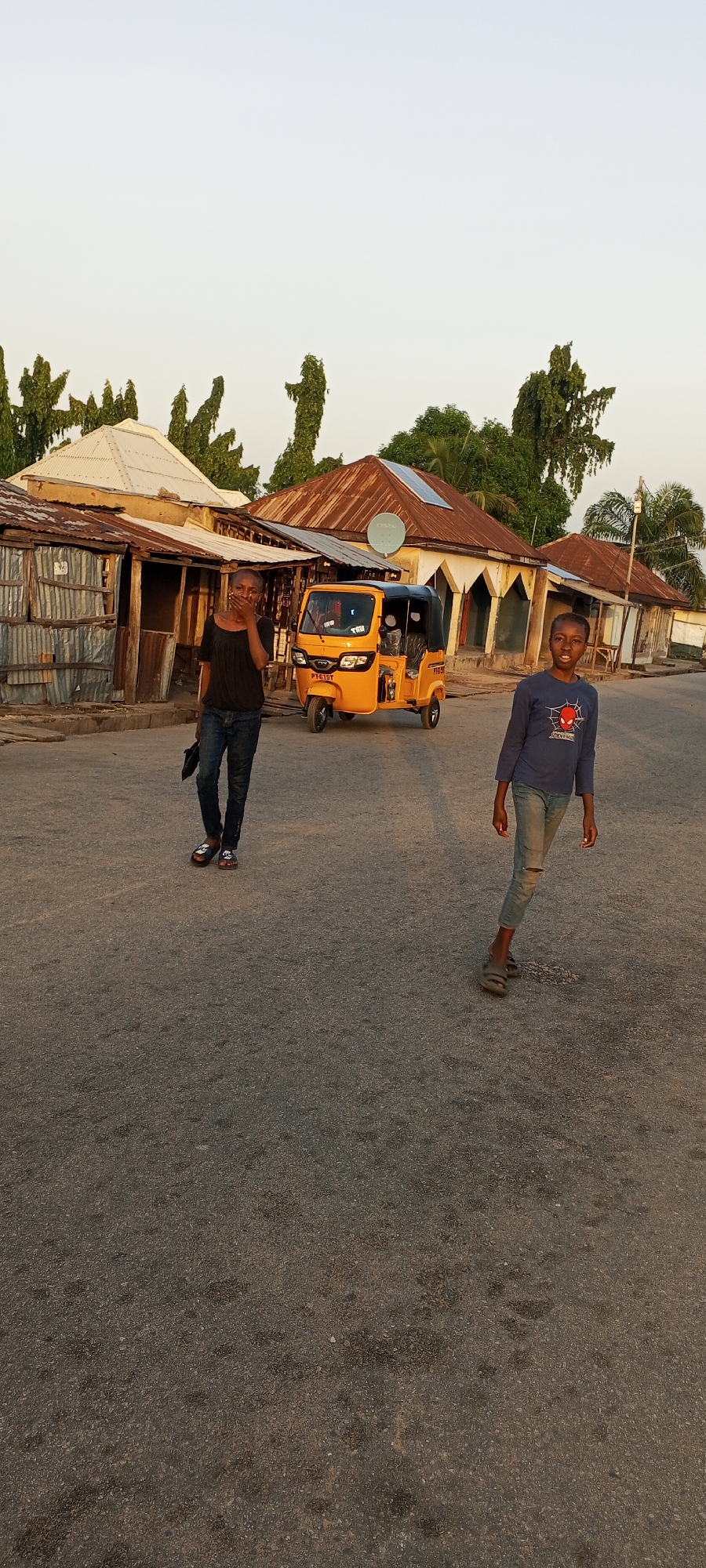 In Jalingo, Nigeria, a street scene unfolds during what appears to be late afternoon. The paved road is empty of vehicles except for a bright yellow auto-rickshaw, identified by license plate "PT-610T", parked on the right side of the street. Behind the auto-rickshaw, a row of single-story buildings lines the street, characterized by corrugated metal and rusty tin roofs, with some showing signs of wear. Trees with lush green foliage are visible behind the buildings, contributing to a rustic environment. Two children are present on the street. On the left, a child dressed in a black t-shirt and blue jeans stands with their hand covering their mouth, holding a black object in their other hand. On the right, another child, wearing a blue t-shirt with a graphic print and distressed blue jeans, is walking and looking towards the camera. The overall impression is of a casual, everyday moment in a Nigerian urban setting.