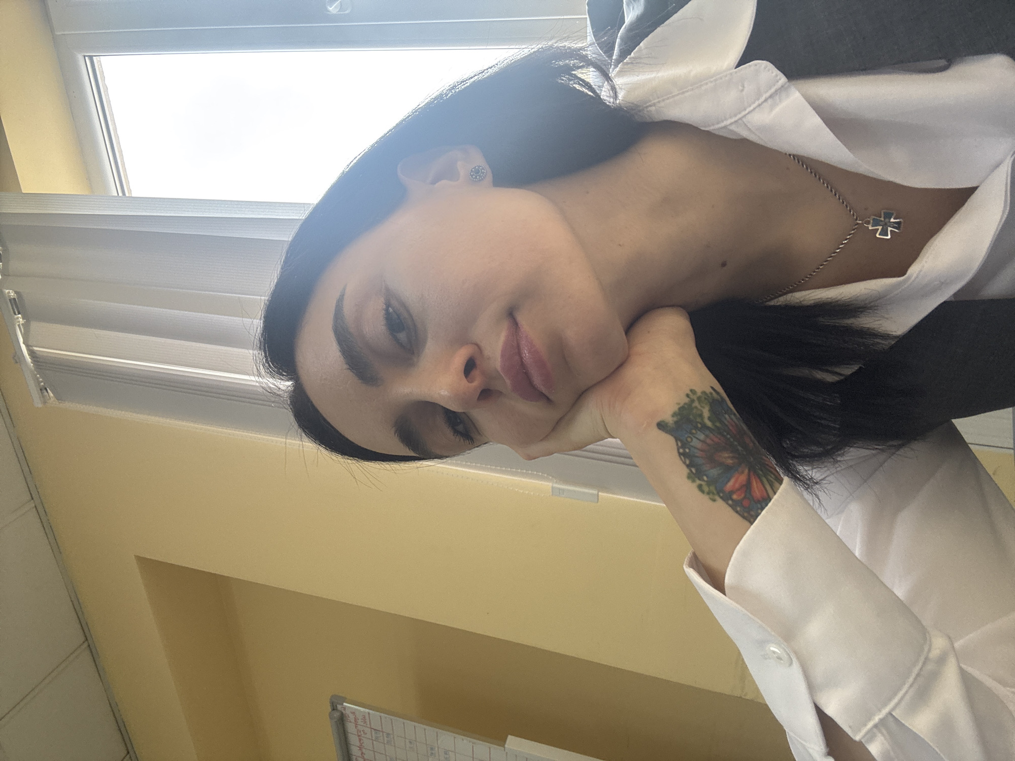 A woman rests her chin on her hand. She has dark hair and wears a white collared shirt with a silver necklace bearing a cross pendant. There is a butterfly tattoo on her wrist. She is indoors, possibly in an office, as suggested by the blinds on the window and a whiteboard visible in the background. The yellow wall and the woman's appearance could indicate the image was taken in Tbilisi, Georgia.