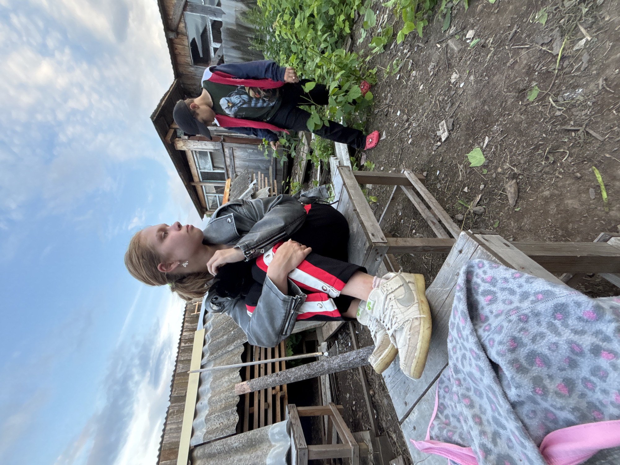 This image depicts two individuals in an outdoor setting that appears to be a rural or dilapidated area, possibly in Маркидоновка, Russia. The scene is characterized by weathered wooden structures, overgrown vegetation, and a cloudy sky, suggesting it might be daytime with overcast weather.
In the foreground, a young person, likely female given the long hair and earrings, is seated on a wooden structure. She is wearing a black leather jacket, black pants with red and white stripes, and light-colored sneakers. Her gaze is directed upwards and to the right, and her posture suggests a relaxed or contemplative mood.
Behind and above her, another individual, likely male, is standing. He is wearing a black baseball cap, a dark jacket over a graphic t-shirt with a bird or owl design, and black pants. He is also wearing red slip-on shoes. He is looking downwards towards the ground.
The overall impression is one of a casual, perhaps slightly neglected, outdoor environment. The wooden structures could be part of an old building or fence. The presence of a rake leaning against a wooden beam suggests some form of outdoor activity or maintenance. The image captures a moment of pause or interaction between the two individuals in this rustic location.