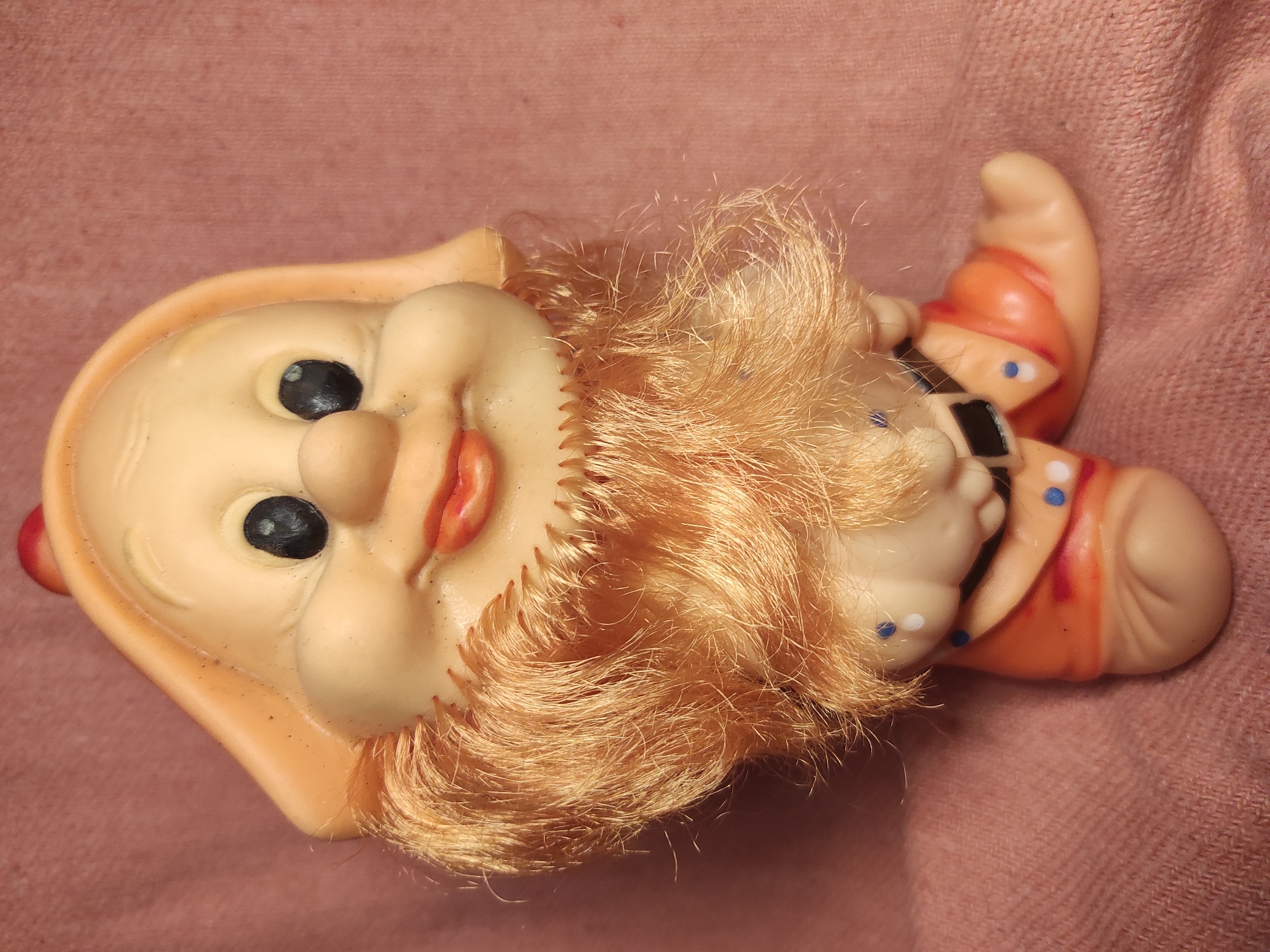 This is a close-up, overhead shot of a vintage rubber doll lying on a pink, textured surface. The doll appears to be a gnome or a similar fantasy creature, with a large, rounded head and a smiling face. Its skin is a pale peach color, with rosy cheeks. It has prominent black eyes, a bulbous nose, and red lips. A fringe of bright orange, synthetic hair frames its face and extends down its neck, resembling a beard. The doll is wearing an orange outfit with blue and white polka dots and black trim, suggesting a playful or whimsical character. The setting is simple, with the doll being the sole focus against the plain pink background, possibly a fabric surface like a blanket or tablecloth. There are no discernible location cues, time of day indicators, or weather information. The overall emotion conveyed by the doll's expression is one of cheerful innocence. No visible text is present in the image.