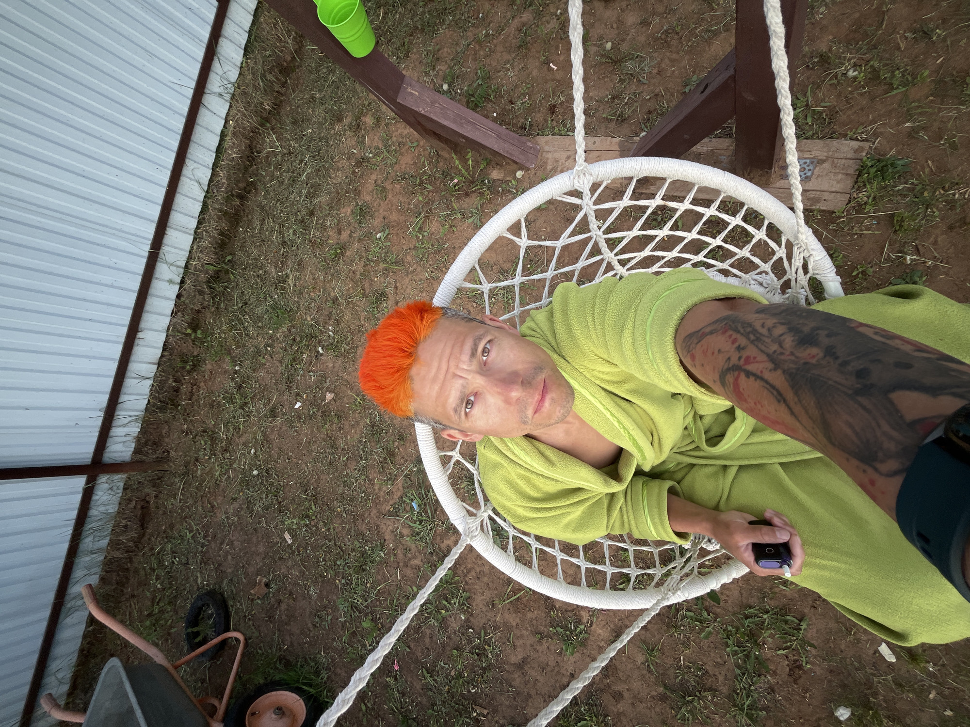 The image shows a man sitting in a white macrame swing chair outdoors. The man has orange hair and is wearing a light green robe. He is looking at the camera and holding a black object in his right hand. His left arm has a large tattoo. The swing is suspended from a wooden structure. In the background, there's a corrugated metal wall on the left and grassy ground with a wheelbarrow at the lower left corner. A green cup sits on a wooden surface. The location is Chaikovsky, Russia.
