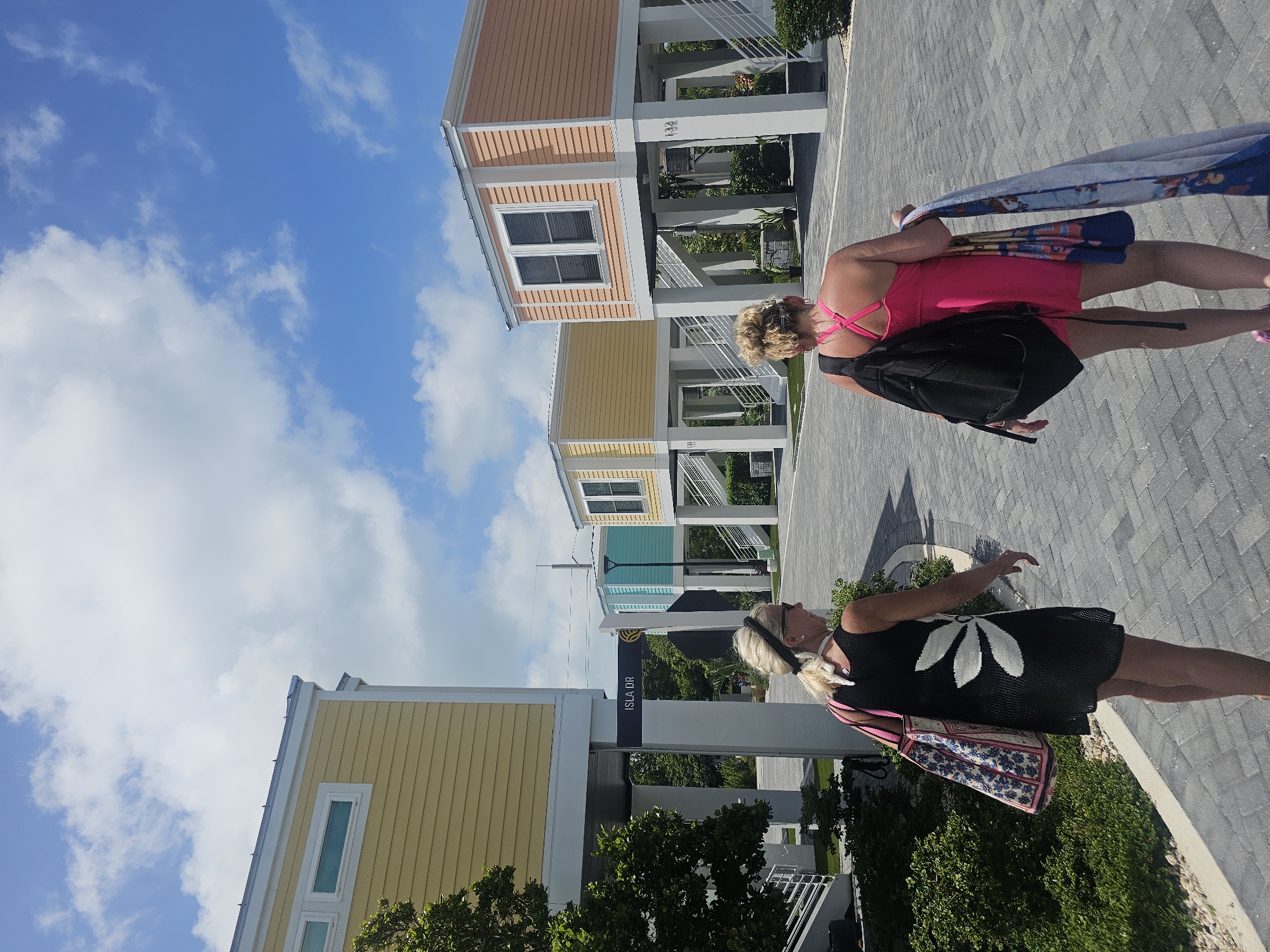 The image shows two women walking on a paved walkway in Islamorada, Florida, United States of America. The women are walking away from the camera, one wearing a pink dress and carrying a black backpack and a folded blue and white towel. The other woman wears a black dress with a large white floral design and carries a patterned bag. The background features a row of colorful, stilted houses with visible numbering on each house. The sky is blue with scattered white clouds. A sign labeled "Isla Dr." is visible near the first house. Lush greenery is present along the walkway.