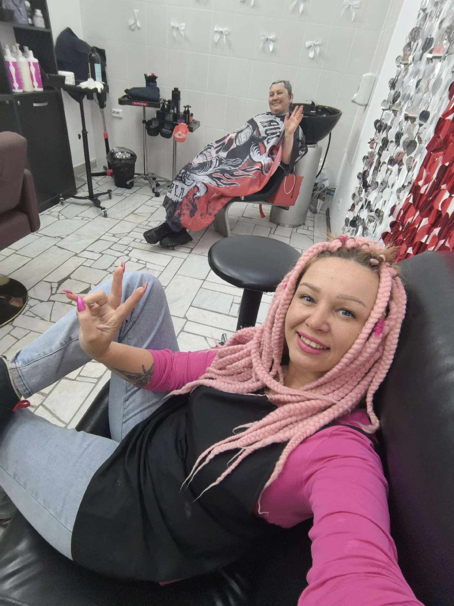 In a brightly lit room, possibly a hair salon in the city of Chaikovsky, Russia, a woman with pink braids is reclining in a black chair. She is wearing a pink long-sleeved shirt under a black salon cape and light blue jeans. She is smiling and holding up her hand, displaying long, pink fingernails. In the background, another woman sits in a salon chair with a cape over her shoulders, waving. The environment includes salon equipment, white tiled walls decorated with bows, and a silver and red decorative curtain. The floor is covered in light-colored tiles.