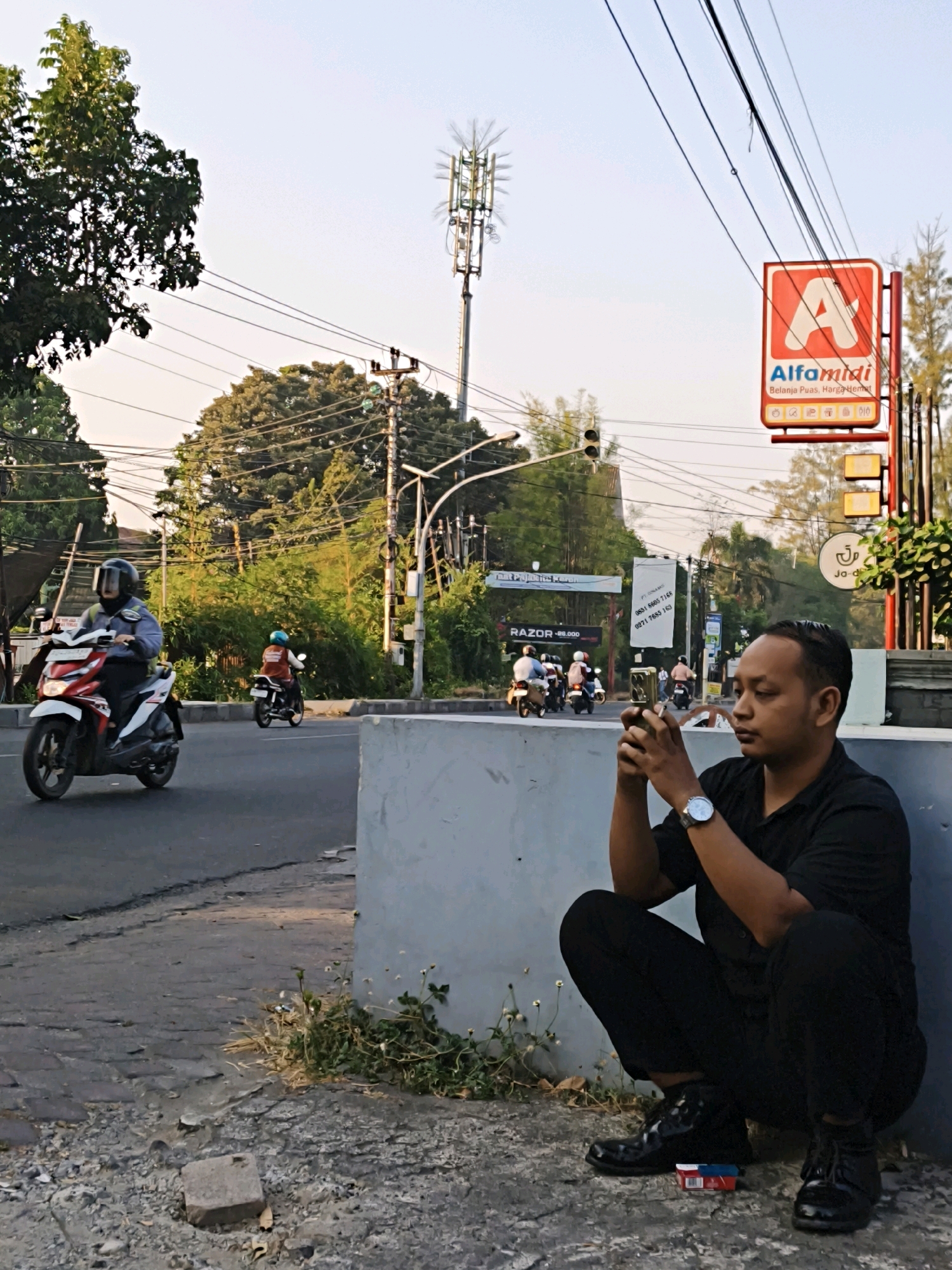 The image depicts a street scene in Surakarta, Indonesia, during daylight hours. In the foreground, a man is crouched down on the sidewalk, looking at his mobile phone. He is wearing a black short-sleeved shirt, black pants, and black shoes. A pack of cigarettes is placed on the ground near his feet. In the middle ground, motorcycles are driving on the road. One motorcycle in the left lane is being ridden by a person wearing a helmet and a blue jacket. Another motorcycle in the right lane is ridden by a person wearing a helmet and an orange vest. The background shows a busy street with buildings, trees, traffic lights, and a tall telecommunications tower. An Alfamidi store sign is visible on the right side of the image. The overall environment suggests an urban setting.