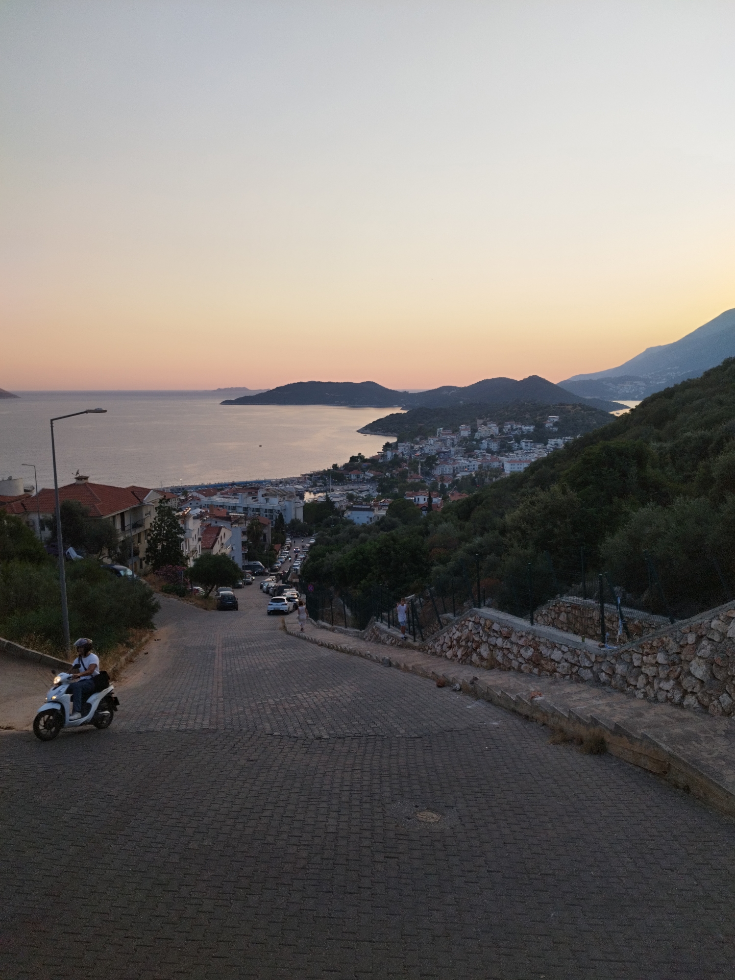 This image captures a descending cobblestone street in Kaş, Turkey, overlooking the Aegean Sea and the town. The sky is a gradient of pale blue to soft orange, indicating either sunrise or sunset. In the foreground, a person rides a white scooter downhill. The street is lined with trees and a stone wall. Further down the street, several cars are parked or in motion, and buildings with red-tiled roofs are visible, leading towards the harbor. The sea is calm, with distant islands and hills forming the horizon. The overall impression is of a quiet, picturesque Mediterranean town.