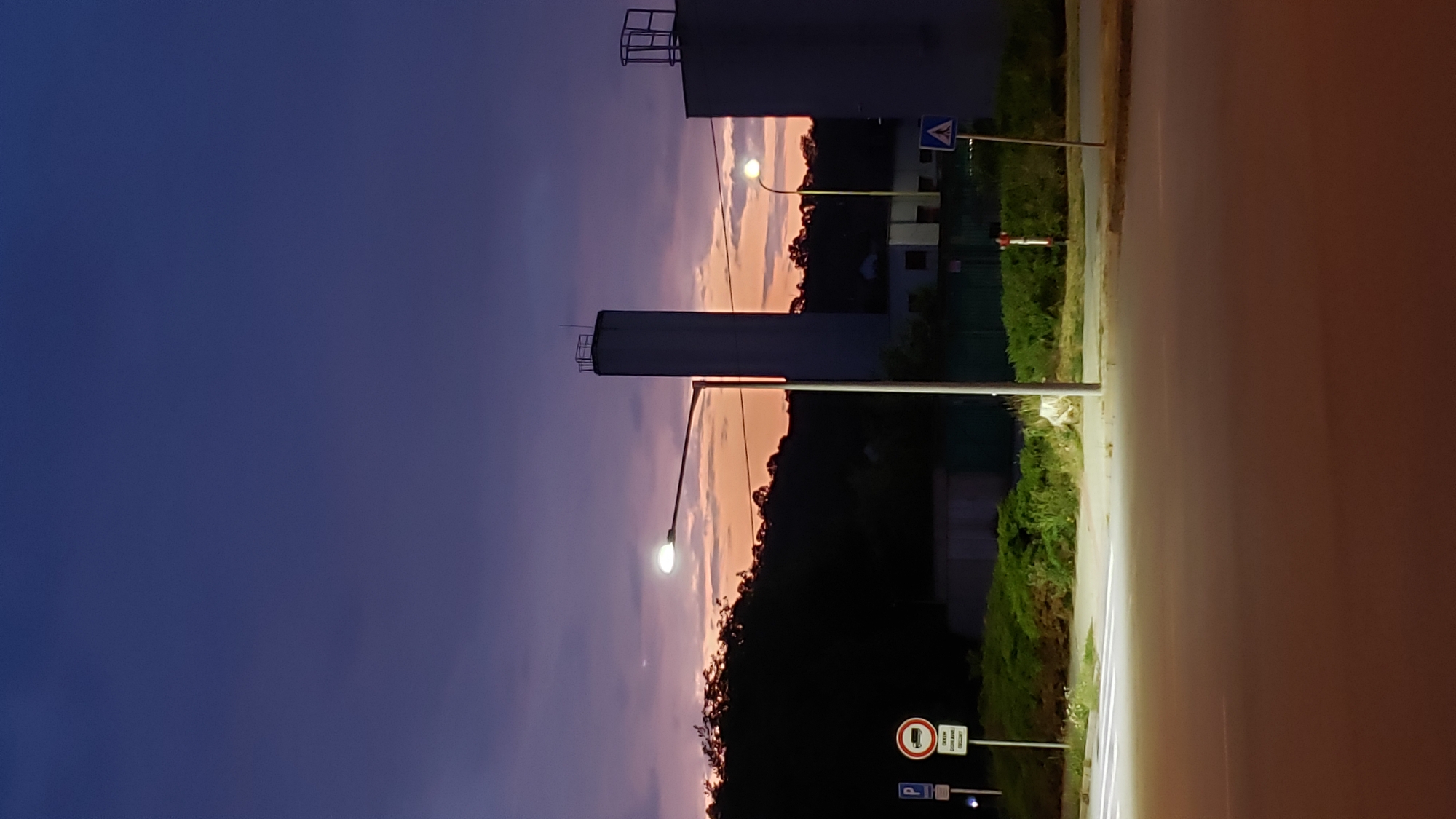 The image depicts a twilight scene in Prešov, Slovakia, likely near an industrial or infrastructure area. In the foreground, a crosswalk is illuminated by a bright street lamp, with an asphalt road stretching out in front. To the left of the crosswalk, a parking sign and a traffic sign prohibiting trucks are visible. Beyond the road, a line of dark trees forms a silhouette against the colorful sky.
Dominating the background are two tall, cylindrical structures, possibly part of an industrial facility or utility plant. The left structure is a white silo or chimney, while the structure to its right is a grey building with scaffolding. A second, smaller street lamp is visible to the right of the silo, also illuminated. The sky is a gradient of deep purple and orange, indicating sunset or sunrise. A lone white dog is visible near the base of the illuminated street lamp on the left.