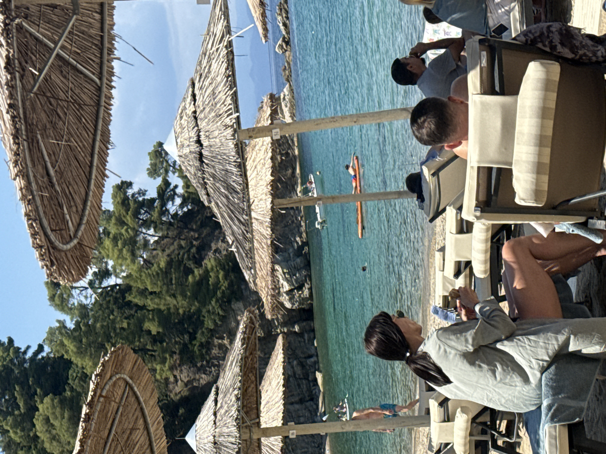 The image depicts a sunny day at a beach in Sithonia Municipal Unit, Greece. In the foreground, several people are seated on lounge chairs under thatched umbrellas. One woman, wearing a light green jacket and shorts, is holding a small object in her hands. Behind her, a man is facing away from the camera, also seated on a lounge chair. Further back, a man in a grey t-shirt is visible, looking towards the sea. The sea itself is a clear turquoise color, with a few boats and people visible in the water. One person is on a paddleboard, another is swimming, and a third appears to be on an inflatable. In the background, rocky cliffs covered in green vegetation rise from the water, characteristic of the Sithonia coastline. The sky is blue with a few scattered clouds.
