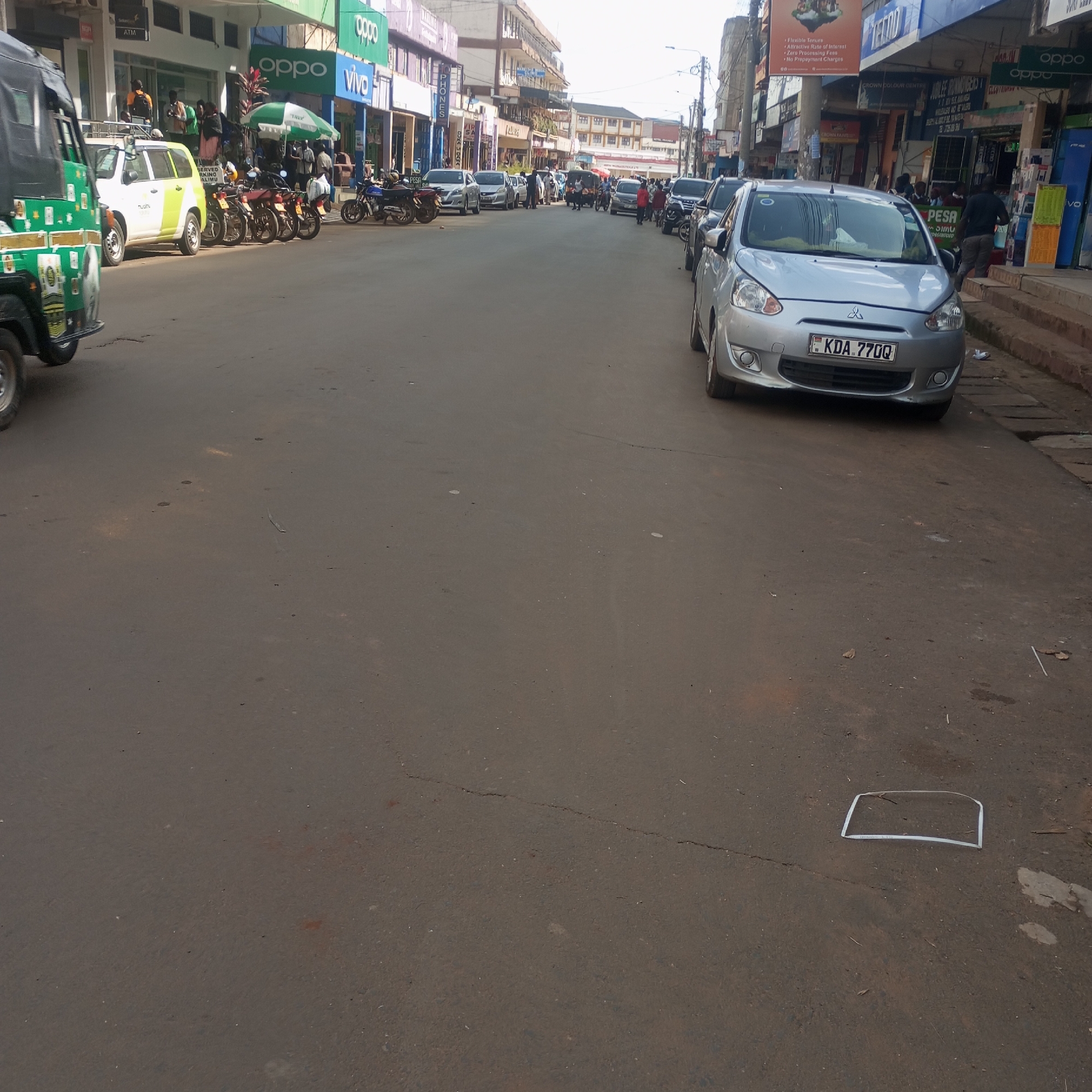 The image depicts a street scene in Sheywe ward, Kenya. The foreground is dominated by a paved road. A silver Mitsubishi Mirage car is parked on the right side of the road, bearing the license plate KDA 770Q. Further down the street, other vehicles, including a green tuk-tuk, cars, and motorcycles, are parked along both sides of the road. Pedestrians are visible on the sidewalks and moving along the street. Businesses line the street, with signs visible for OPPO and VIVO. Street signs and utility poles are also present. A piece of trash is visible on the street surface in the foreground.