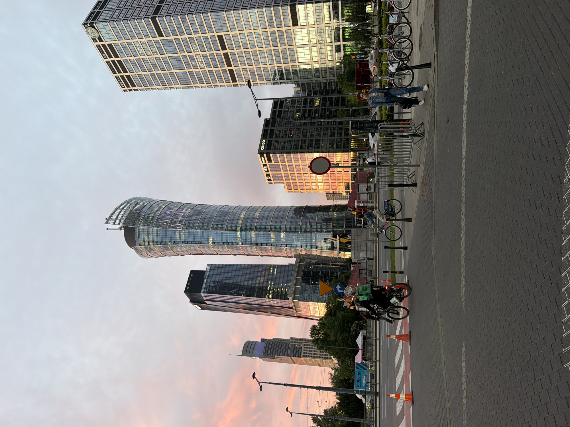The image depicts a street scene with several modern high-rise buildings in the background. The sky is a pale blend of pink and grey, suggesting dawn or dusk. In the foreground, a street paved with grey rectangular blocks runs parallel to the base of the image.
Several tall, contemporary buildings of varying designs dominate the view. From left to right, the buildings are as follows: A tall glass tower with a distinct curve, multiple levels, and a silver-colored top; a dark, rectangular skyscraper with windows; another skyscraper with an angular design; and a taller glass building with a grid-like pattern of windows.
Along the right edge of the image, parked bicycles are lined up and attached to a metal rack. Nearer the street, a pedestrian wearing a denim jacket and jeans stands beside the bicycle rack. A bicycle rider in dark clothing with a green delivery backpack passes by on the street. Orange traffic cones are placed along the edge of a pedestrian crossing.
Streetlights, signs, and other street furniture are also visible.