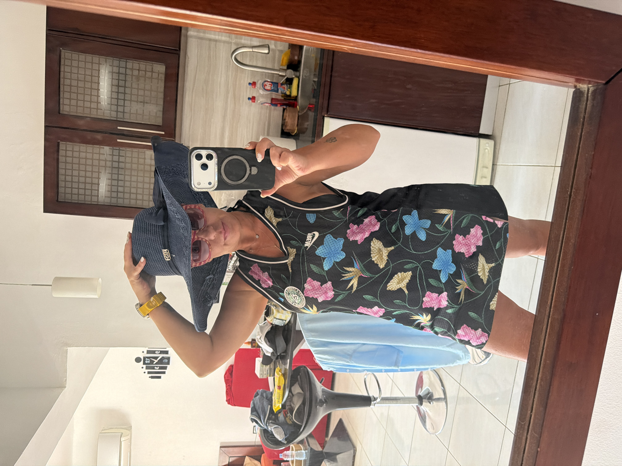 A woman stands in a room, possibly an apartment or hotel, in Sharm El Sheikh, Egypt, taking a mirror selfie. She is wearing a dark blue wide-brimmed hat, pink sunglasses, and a black tank top with a floral print. Her left hand is on top of her hat, and her right hand holds a smartphone, also visible in the reflection. The room appears to be a living area or bedroom with tiled floors and wooden cabinets. In the background, a red sofa and a bar stool with items on it are visible. The overall lighting suggests it is daytime.