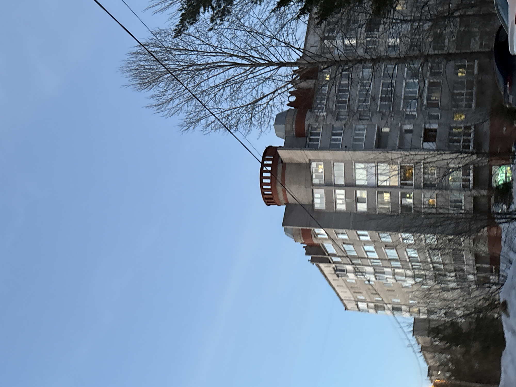 The image shows a multi-story apartment building in Berezniki, Russia. The building has a distinct cylindrical feature with a red railing near the top. The architecture is typical of Soviet-era construction. The sky is a clear blue. Bare tree branches are visible in the foreground and partially obscure the building. Power lines run across the top of the frame. Patches of snow are visible on the ground in the lower right corner.