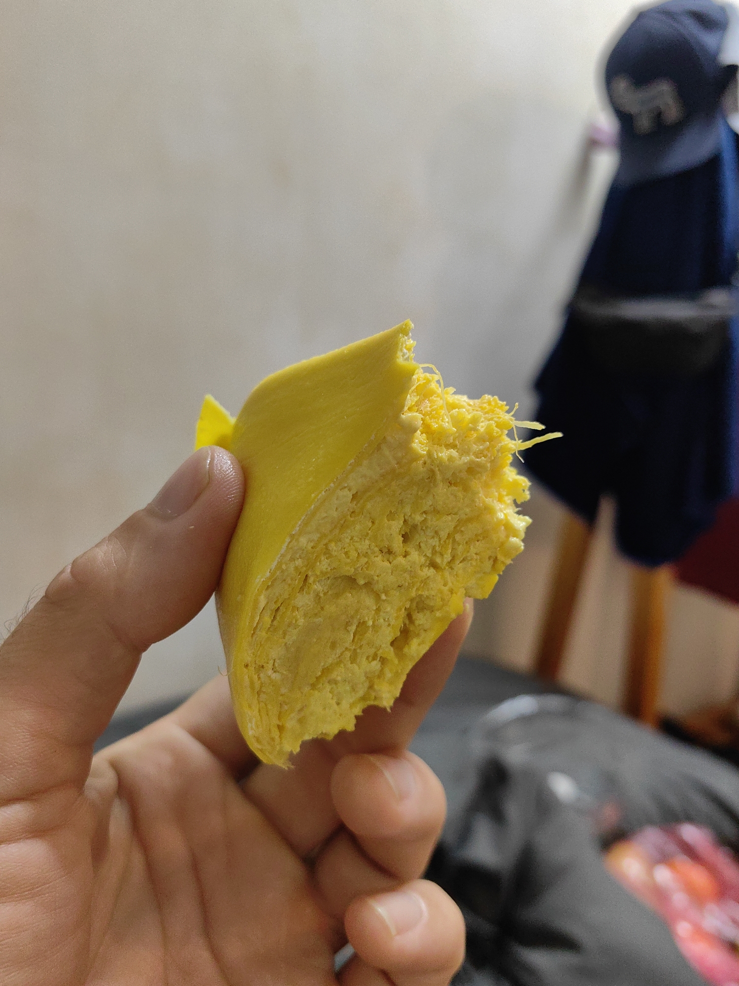 A hand holds a chunk of bright yellow durian fruit indoors. Background objects include a white wall, a dark jacket hanging on a chair, and a baseball cap. The location is most likely in Thành phố Hà Giang, Vietnam, as that was provided with the prompt.