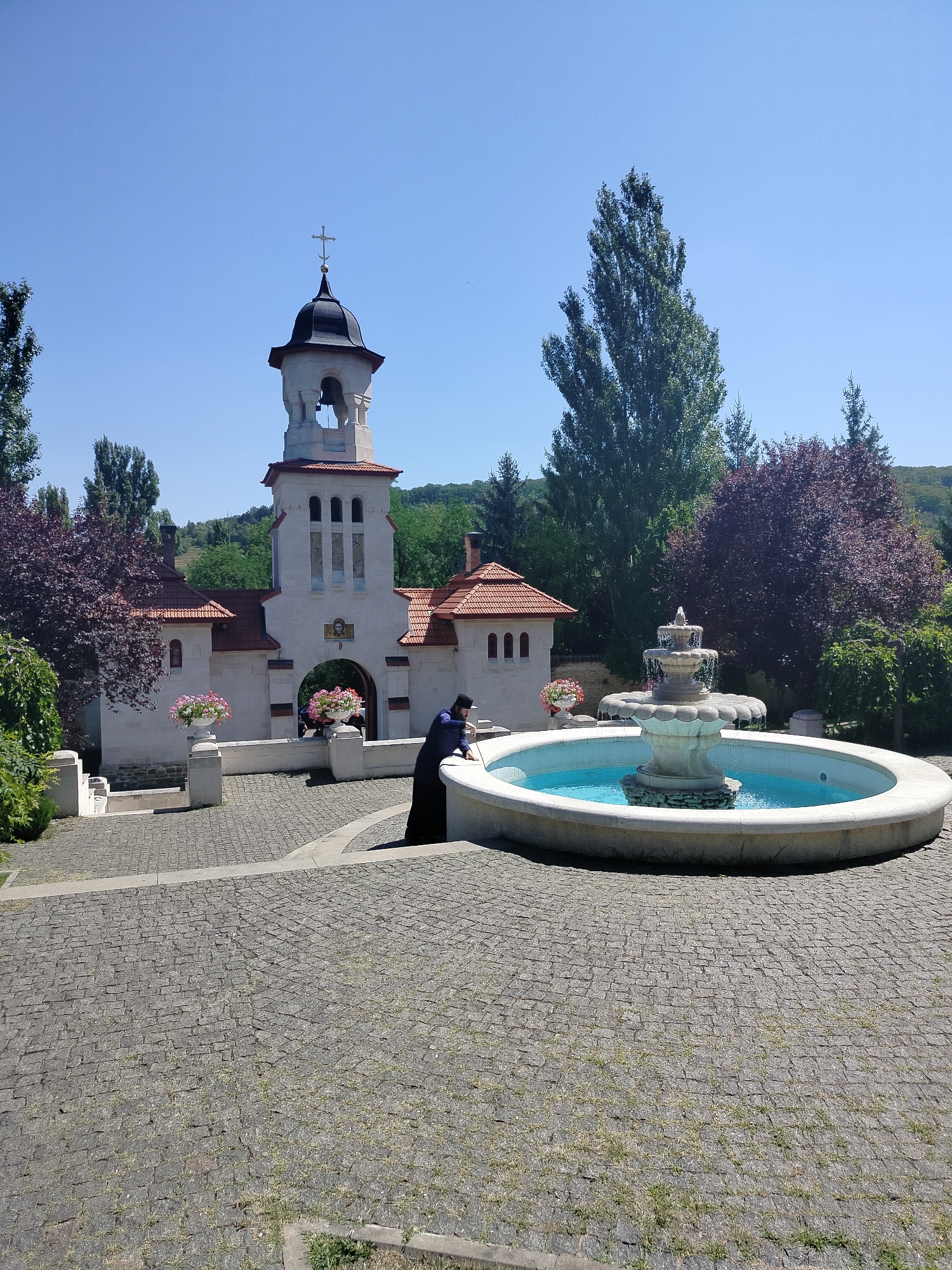 A priest in a dark robe is seen near a fountain in front of a church. The church has a bell tower and a cross on top. The fountain is large and round with a multi-tiered centerpiece. The ground is paved with cobblestones. Trees and a clear blue sky form the background.