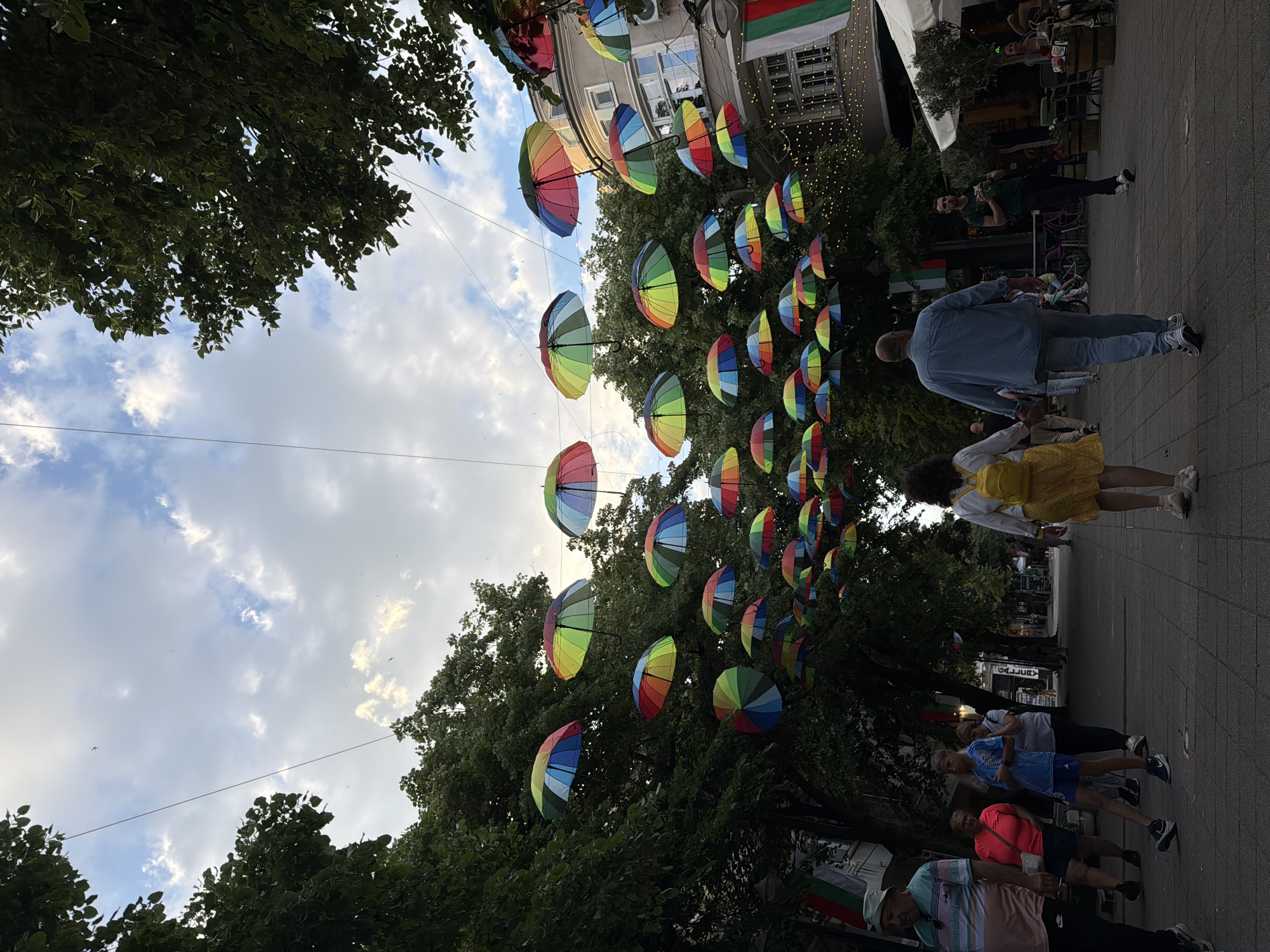 The image shows a street scene in Burgas, Bulgaria. Rainbow-colored umbrellas are suspended above a pedestrian walkway lined with trees. People are walking along the walkway, including a person in a denim jacket, a person in a yellow dress, and a group of people in the foreground. Bulgarian flags are visible. Buildings are in the background. The sky is cloudy with visible sunshine.