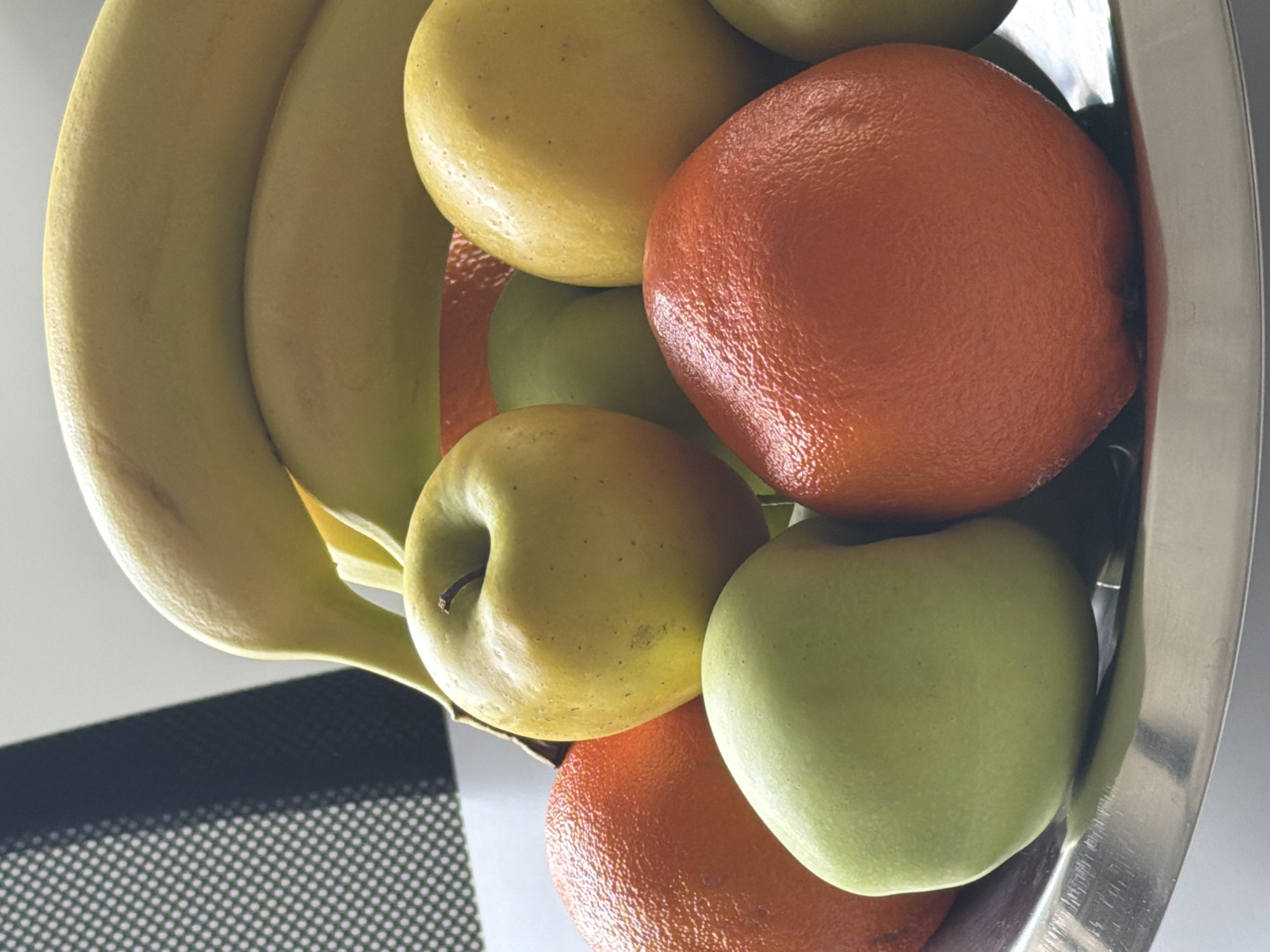 A close-up, high-angle shot depicts a silver bowl overflowing with fruit. Predominantly, two bananas curve along the top edge. Within the bowl, a mix of fruits includes two oranges, and three apples (two green, one yellow). A black, patterned surface is visible in the lower-left corner. No information can be extracted from the image that pertains to the location within Yerevan, Armenia.