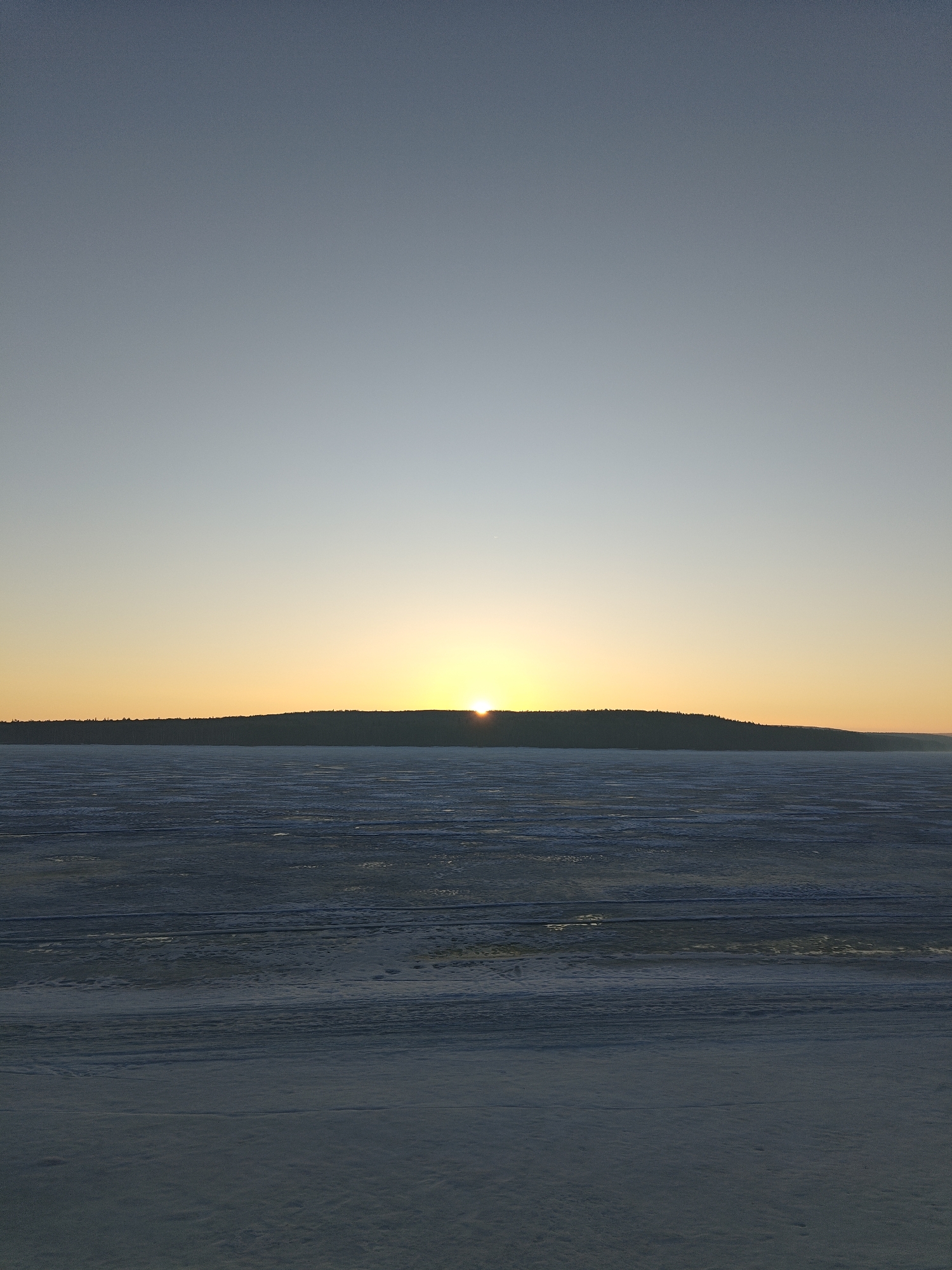 The image depicts a winter sunrise over the Kama River in Chaikovsky, Russia. The horizon is dominated by a dense forest silhouette, with the sun just peeking over the treeline, casting a warm orange glow. The foreground shows a frozen river surface covered in snow and ice, with subtle lines indicating variations in the ice formation. The sky transitions from a light blue at the top to a pale yellow near the horizon. The overall scene is tranquil and cold.