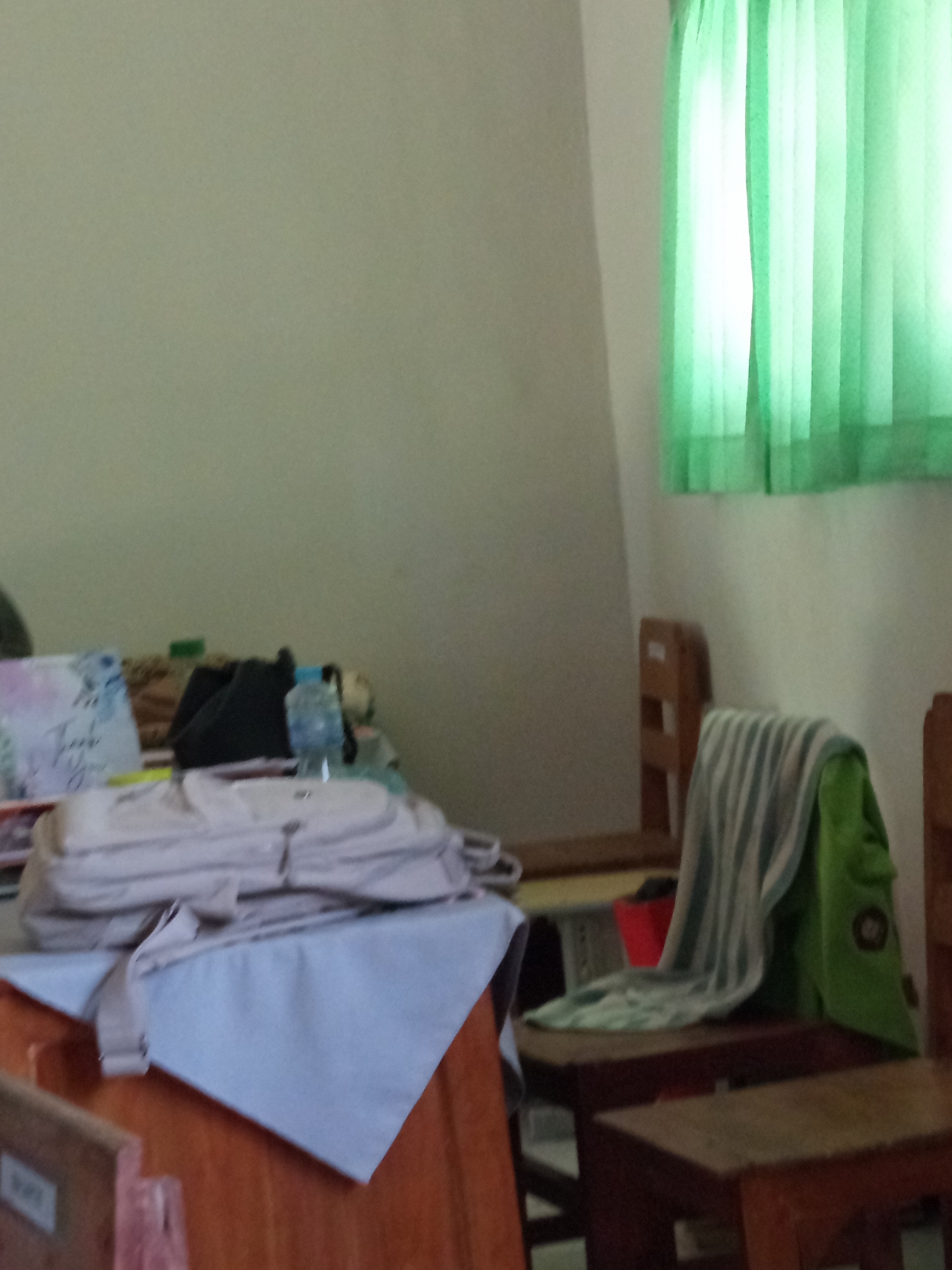 The image depicts a classroom or study area, likely in Lumutan, Indonesia. On a table in the foreground, a light-colored backpack is laid out, partially covered by a light blue cloth. Next to it are other items including a black bag, a water bottle, and a decorative item with writing. To the right, a wooden chair is present, draped with a green cloth and a striped towel. In the background, a light green curtain is visible. The overall impression is one of a functional, perhaps slightly cluttered, space.