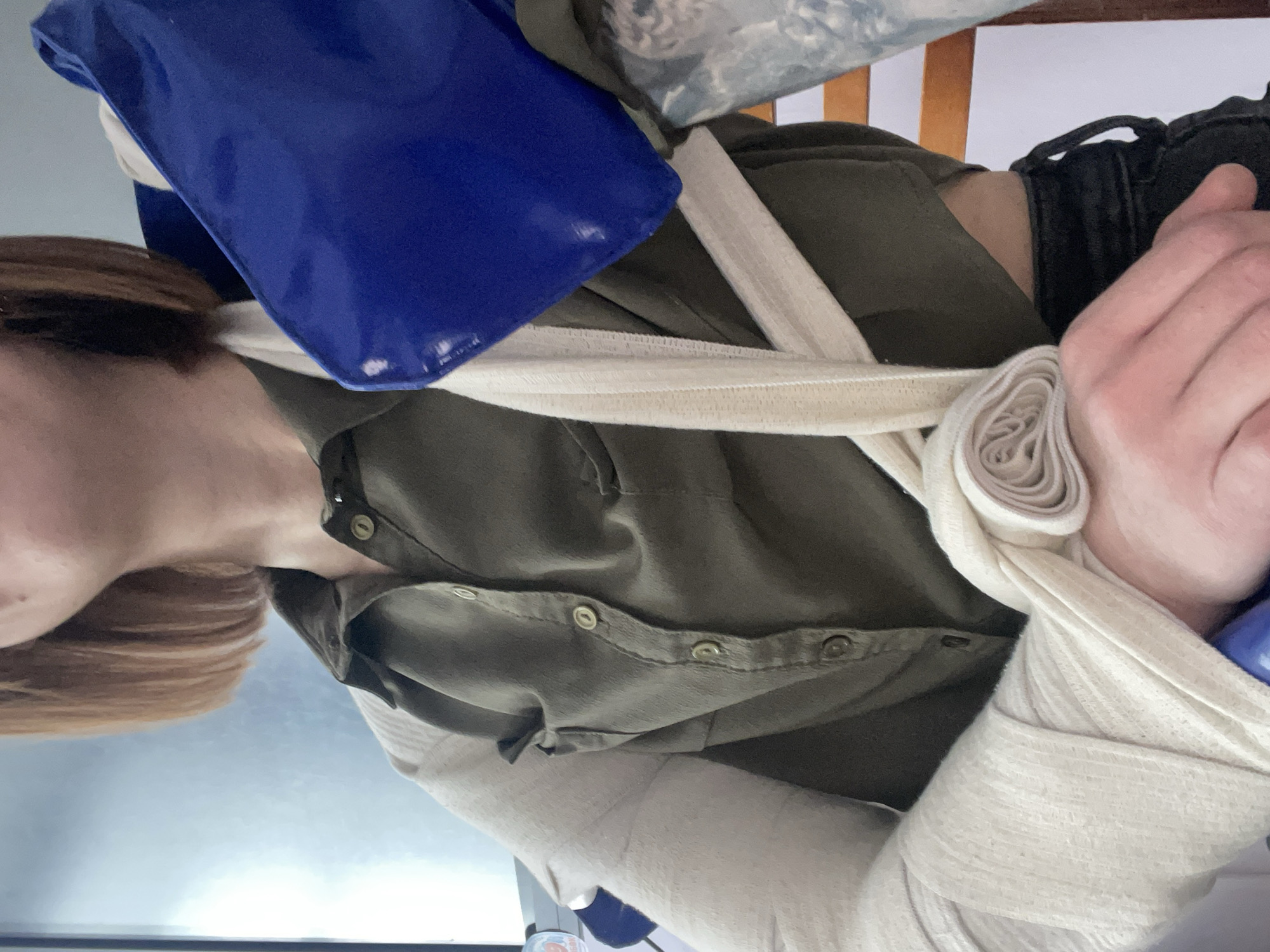 A person with light brown hair is wearing an olive green button-down shirt and black pants. Their arm is supported by a white cloth sling that extends up to their neck, where a blue cold pack rests. The location is not discernable from this image.