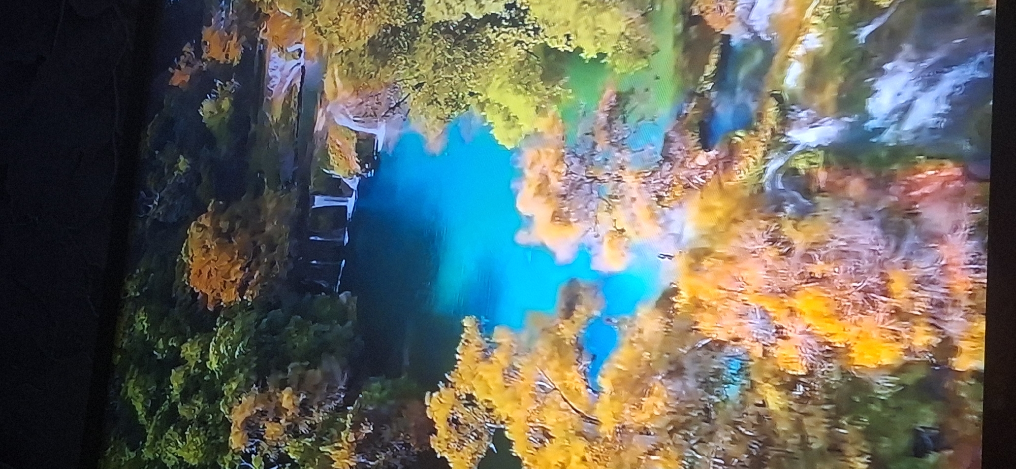 This image shows a vibrant, aerial view of a natural landscape featuring a clear, turquoise body of water surrounded by lush, multi-colored foliage. The water appears to be a lake or a wide river, with several small waterfalls cascading into it from the upper right. The trees exhibit a range of colors, from deep green to bright yellow and orange, suggesting an autumn season or a tropical environment with diverse vegetation. There is a tiny human figure visible standing on a rock formation near the center of the image, appearing to observe the scenery. The overall impression is one of serene natural beauty.
The provided image is an artistic depiction of a natural landscape and does not contain any recognizable landmarks or elements that would allow for its identification within the city of Bila Tserkva, Ukraine. Therefore, no specific location details from Bila Tserkva can be inferred from this image.