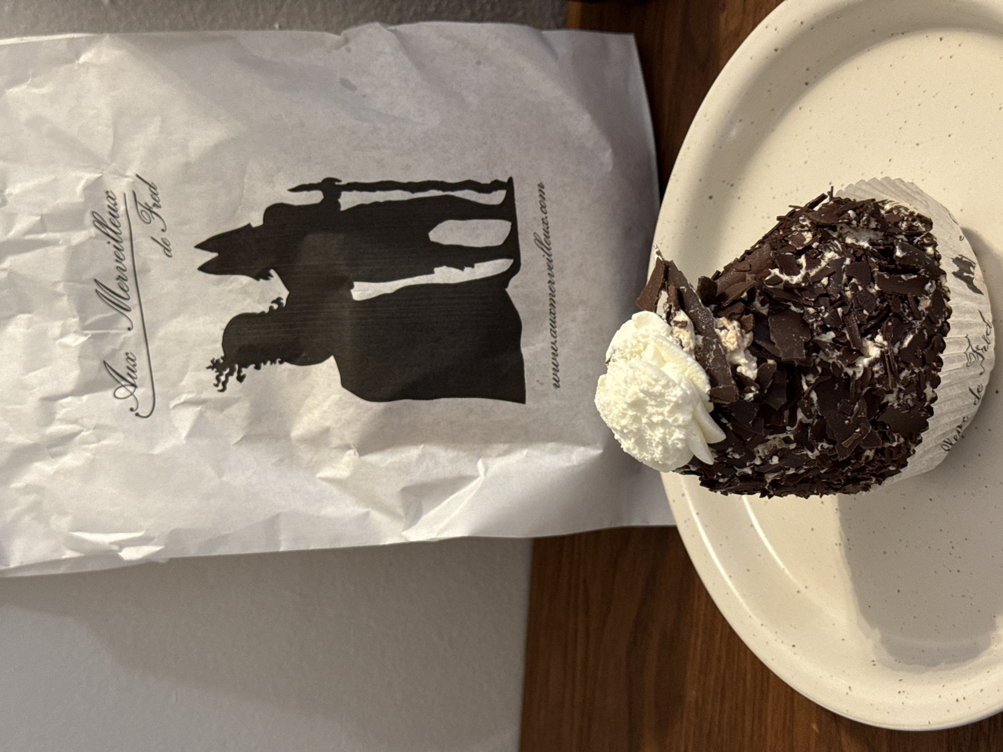 A dessert, likely a "Merveilleux," sits on a speckled ceramic plate next to a white paper bag. The bag is printed with the name "Aux Merveilleux de Fred" and the website "www.auxmerveilleux.com," indicating the dessert likely came from this establishment. The logo on the bag is a black silhouette of people dressed in historical clothing. The dessert consists of a cake base, white cream, and dark chocolate shavings. The scene is on a brown table. Aux Merveilleux de Fred has locations in Montreal, Canada.