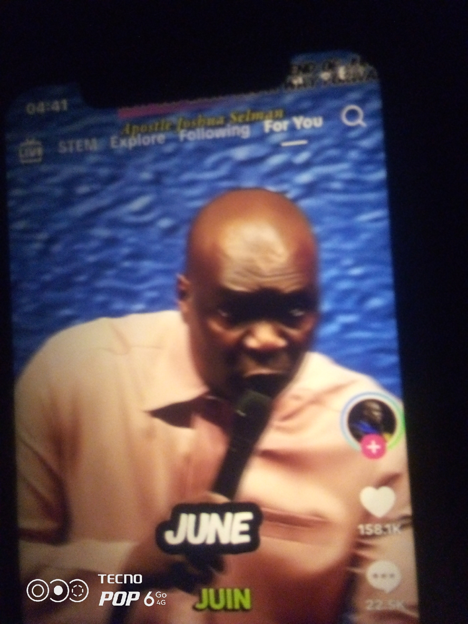 A man with a shaved head and dark skin is shown in a close-up shot, looking directly at the camera with an open mouth. He is wearing a light pink collared shirt. A microphone is held near his mouth. In the background, a textured blue surface is visible. Overlayed on the image are various UI elements typical of a social media app: a timestamp "04:41", text "LIVE STEM Explore Following For You", a search icon, a circular profile picture with a plus sign and a heart icon indicating likes with a count of "158.1K", a speech bubble icon with a count of "22.5K", the word "JUNE" in large white letters, and the text "TECNO POP 6 GO 4G JUIN". The image appears to be a screenshot of a video or live stream on a mobile device, with the phone's branding visible at the bottom left. The content of the video is a presentation or sermon by a speaker. There is no discernible information within the image to indicate the specific location within the city of Howayi, Nigeria.