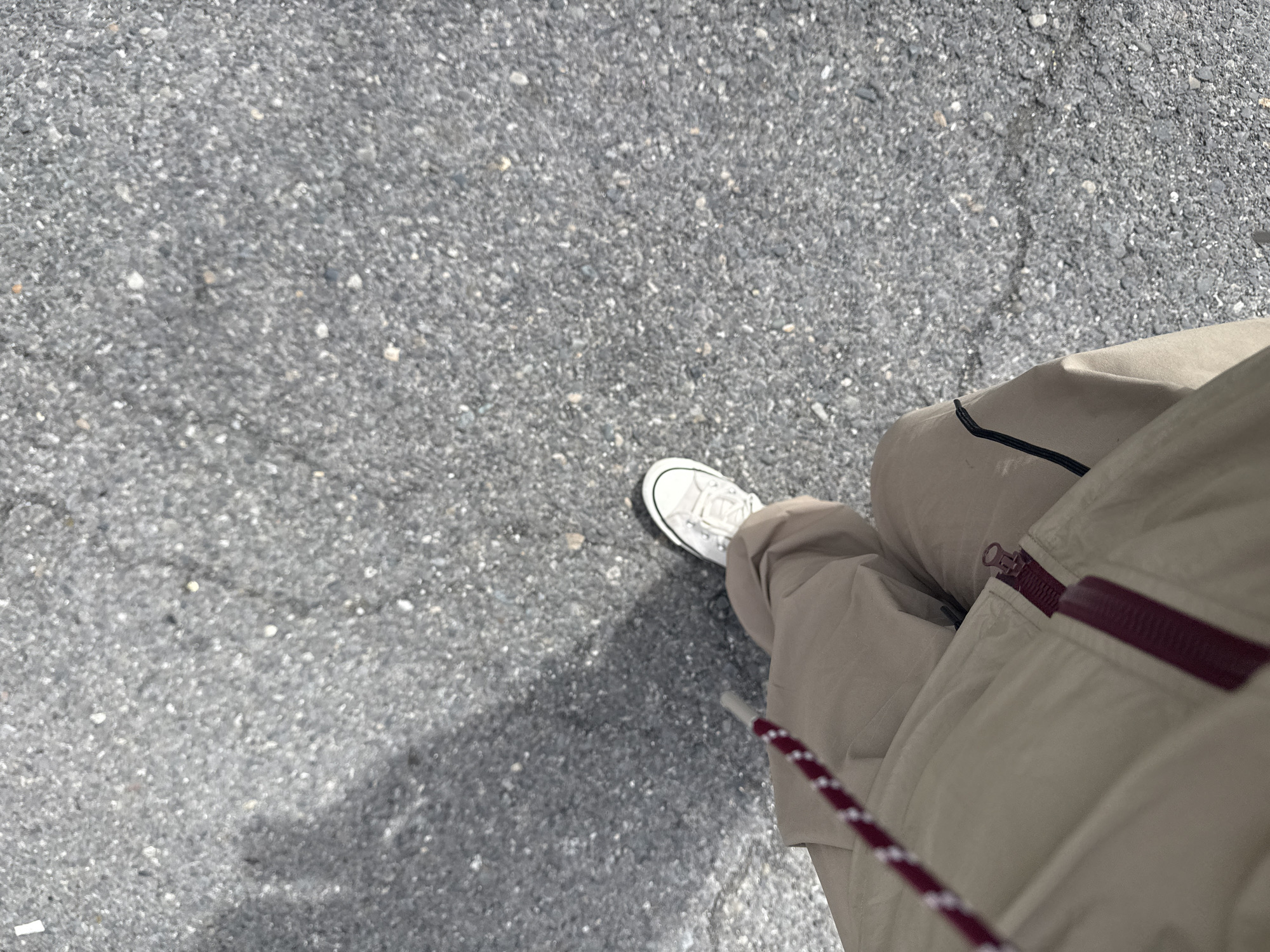 An eye-level, overhead shot depicts a person standing on an asphalt surface. The asphalt, which fills most of the frame, is gray in color and rough in texture. The person is wearing light tan pants with a dark burgundy zipper and trim, and a white sneaker is visible on the left side. A lanyard with a red and white rope is on the left side. The location is an asphalt surface, presumably a sidewalk or a street. The location in the city of Yerevan, Armenia cannot be verified based on the visual information provided.