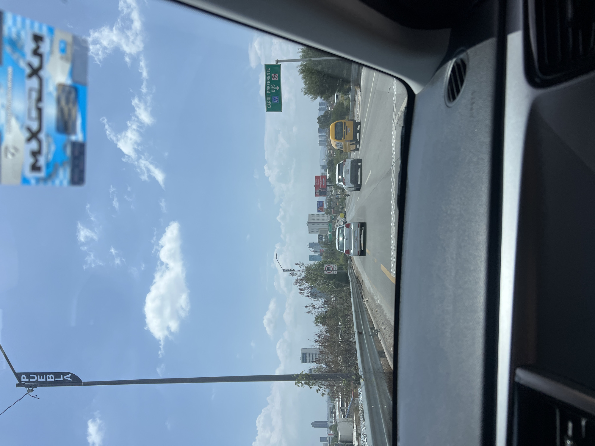 The view from inside a vehicle traveling on a highway approaching San Bernardino Tlaxcalancingo, Mexico. A sign indicates "Carril Preferente Bus 50" (Preferred Bus Lane). There are other vehicles on the highway. In the distance, a city skyline is visible. A sign for "Puebla" is also visible. The sky is blue with scattered clouds.