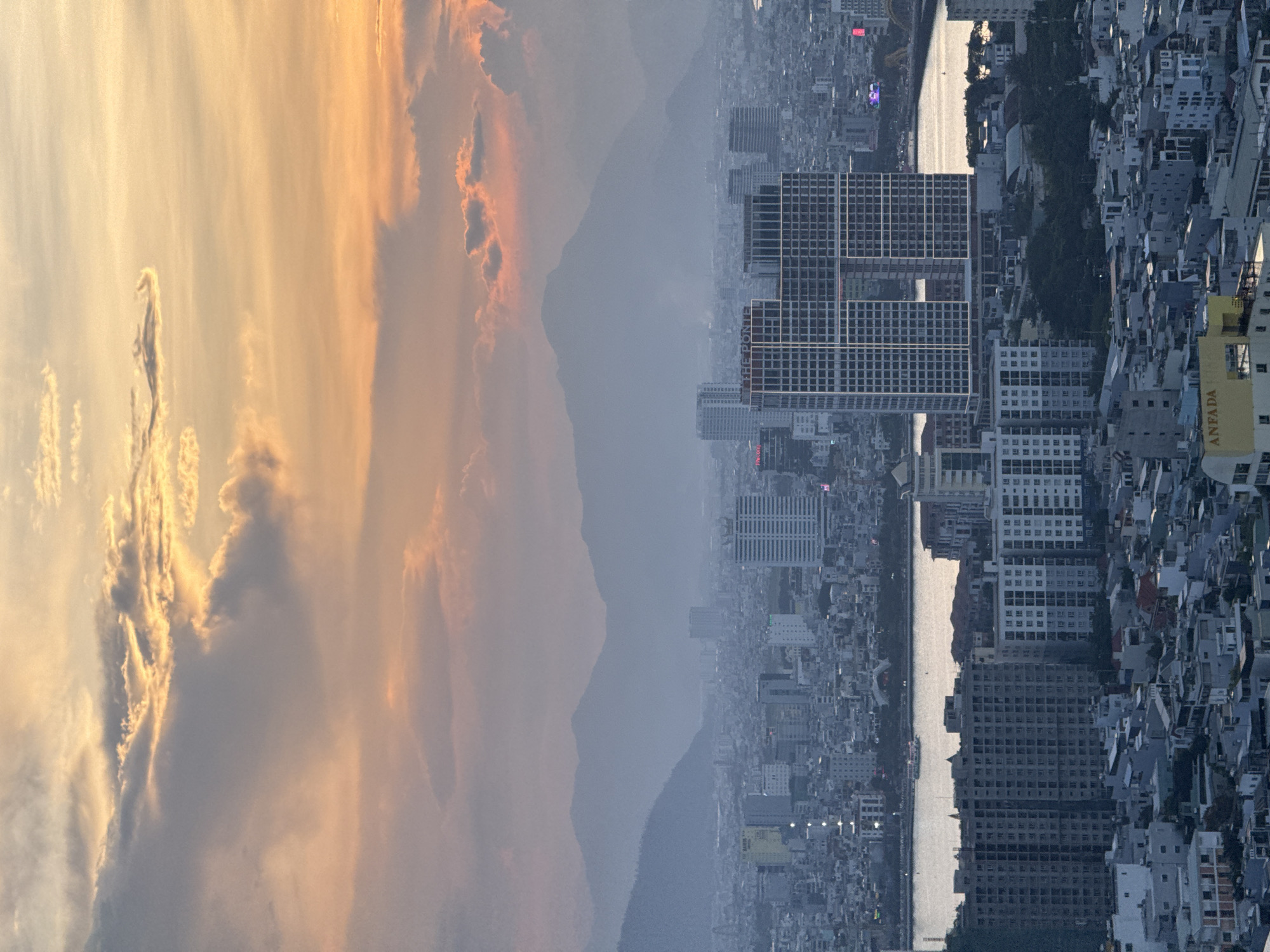 This aerial, vertical photograph depicts a cityscape at dusk, likely from a high vantage point in Đà Nẵng, Vietnam. The sky is filled with dramatic clouds illuminated by the setting sun, casting a warm orange and pink glow. In the distance, hazy mountains form a silhouette against the sky.
The midground features a dense urban environment with numerous buildings of varying heights, interspersed with what appears to be a river or body of water running through the city. Several prominent high-rise buildings stand out, including a modern skyscraper with a distinctive open-section design in the center, and another tall, rectangular building with a grid-like facade. Smaller residential and commercial buildings fill the rest of the urban landscape.
In the foreground, a cluster of lower-rise buildings, characteristic of urban residential areas, are visible. One building in the lower right foreground has signage that reads "ANFADA". The overall impression is one of a sprawling coastal city bathed in the soft, ethereal light of sunset. There are no visible people in the image.