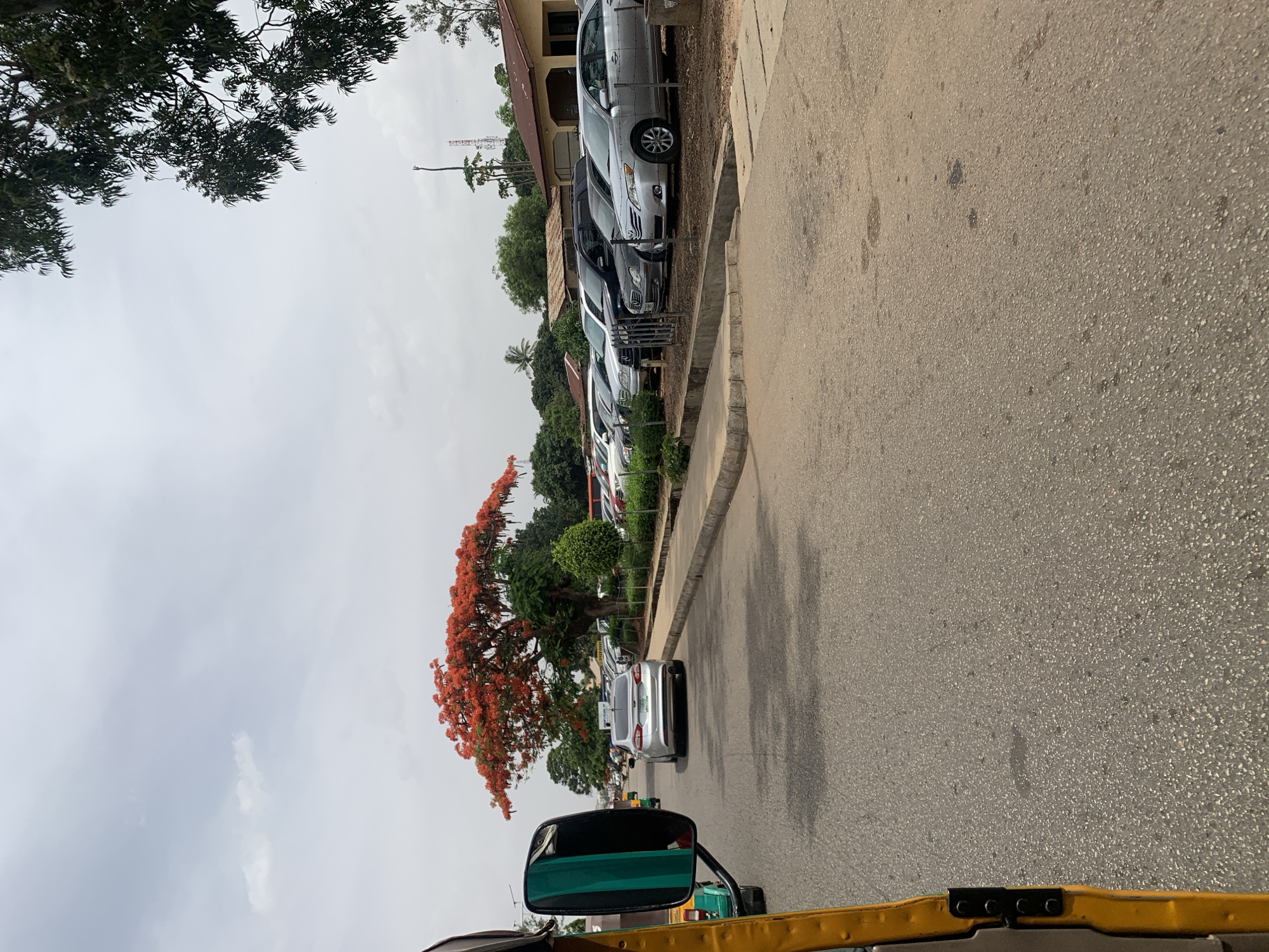 The image shows a street scene in the city of New Stadium, Nigeria. The view is from a moving vehicle, likely a motorized tricycle (keke). A line of parked cars is visible on the left side of the street, adjacent to a curb. A large tree with bright orange flowers is also visible on the left. The street is paved, and the sky is overcast. No people are visible, but the presence of parked cars suggests human activity.