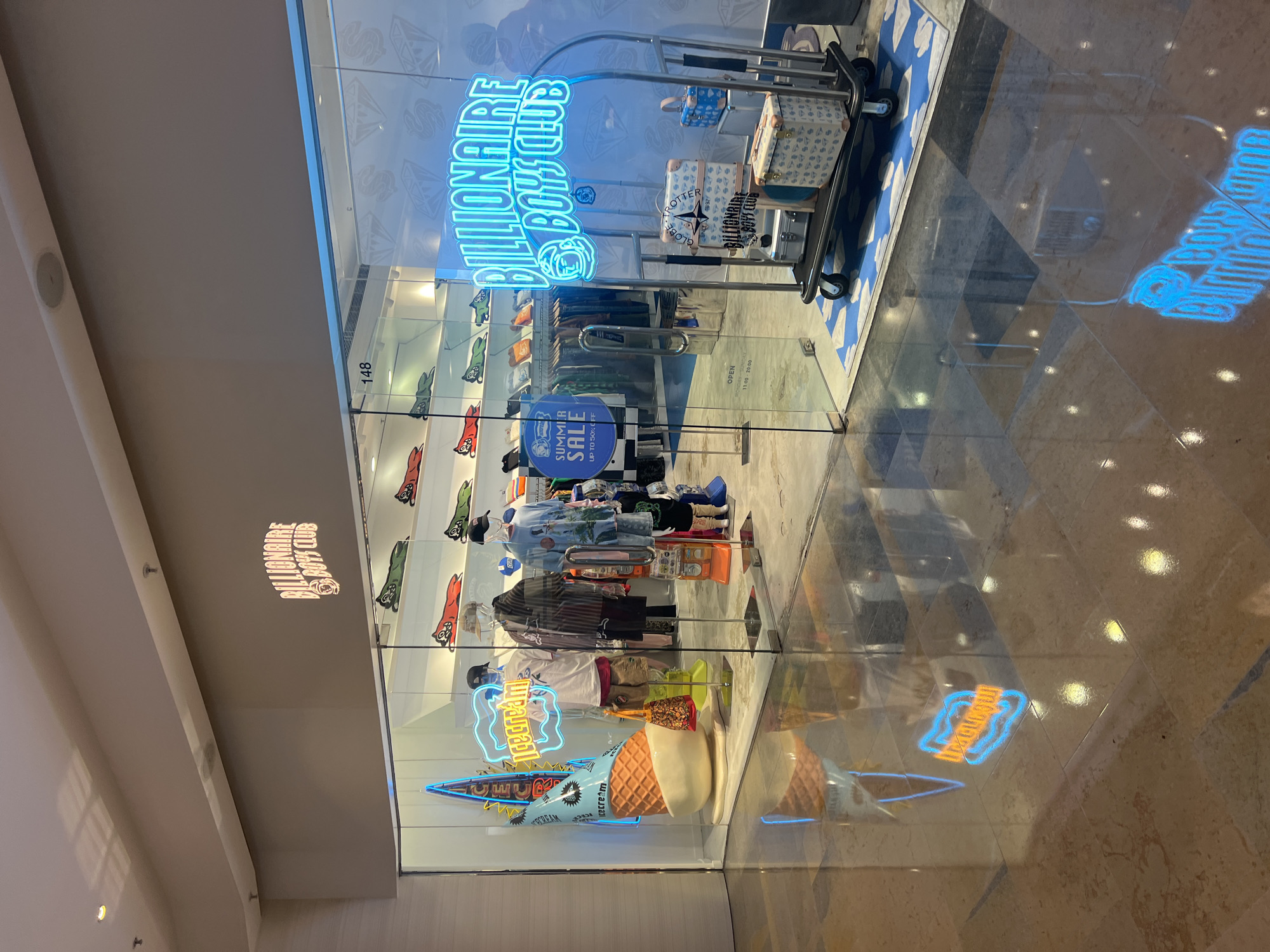 The image shows the storefront of a Billionaire Boys Club store located in Hong Kong, China. The store features a large glass facade with neon signage visible both inside and reflected on the polished floor. Inside, mannequins are displayed wearing clothing from the brand, and shelves are stocked with apparel featuring the brand's signature motifs, such as cartoonish dog-like figures. A prominent ice cream cone sculpture is placed in the window display, along with a neon "icecream" sign. A luggage cart with several suitcases, some branded, is positioned just inside the entrance. The store appears to be open, with a sign indicating summer sale information. The environment is brightly lit, and the reflections on the floor suggest an indoor shopping mall setting. No people are clearly identifiable in the image.