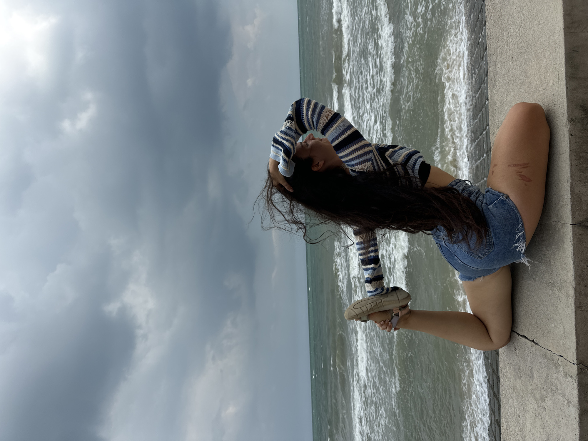 A woman with long, dark hair is performing a yoga pose on a concrete ledge overlooking a choppy, grey sea under a cloudy sky. She is wearing a striped long-sleeved shirt and denim shorts. Her right leg is bent at the knee, with her foot reaching back towards her head, which she is holding with her right hand. Her left leg is bent and tucked beneath her. The environment suggests a coastal location.