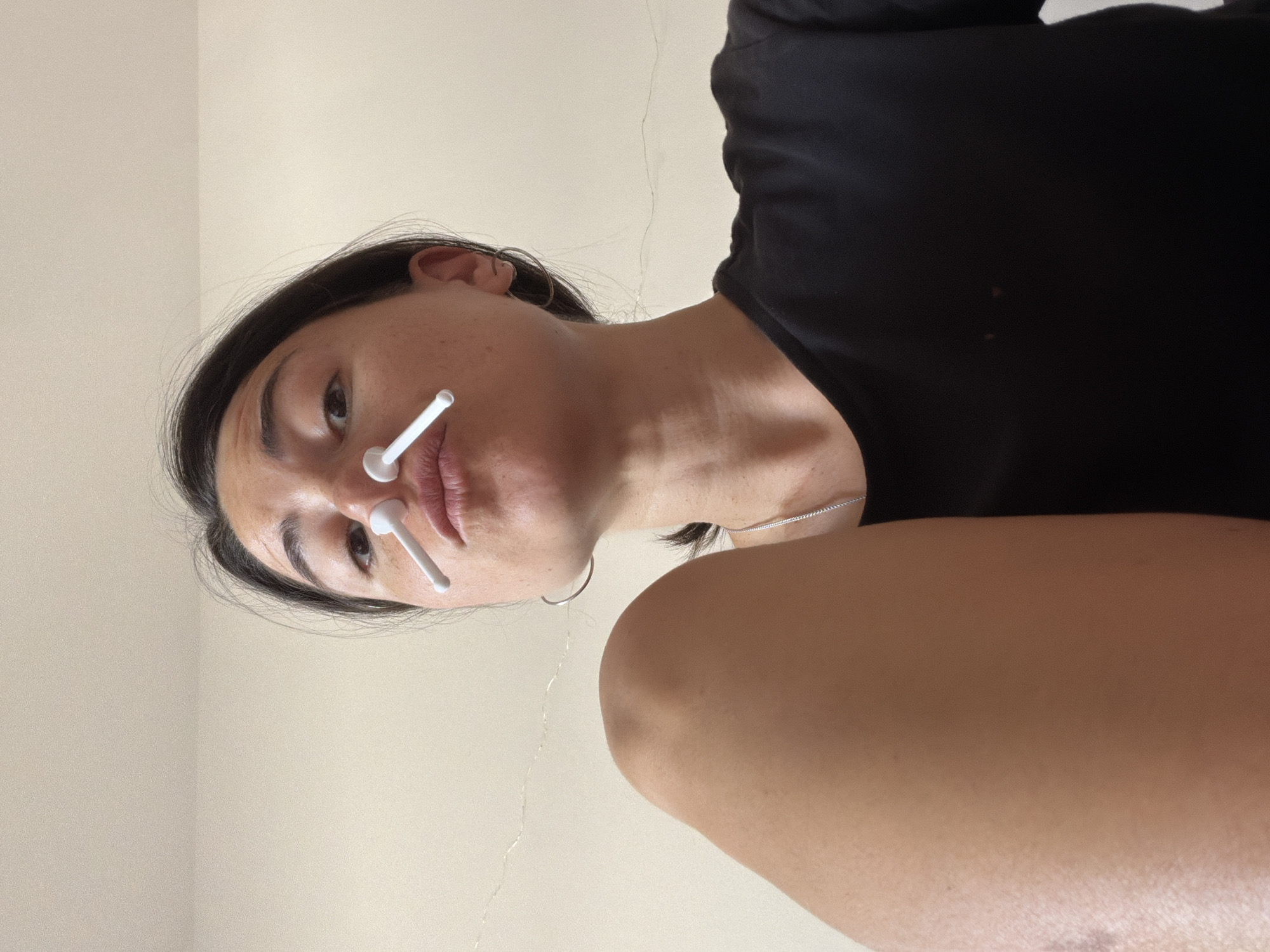 A woman is positioned in the center of the frame, with her face and upper body visible. She is looking down at the camera. Two white plastic objects resembling cotton swabs are inserted into her nostrils. She has dark hair, dark eyes, and is wearing a black top. A silver hoop earring is visible in her left ear. Her skin tone appears light to medium. The background is a plain beige or light-colored wall. The photograph does not provide information about the city of Budva, Montenegro.