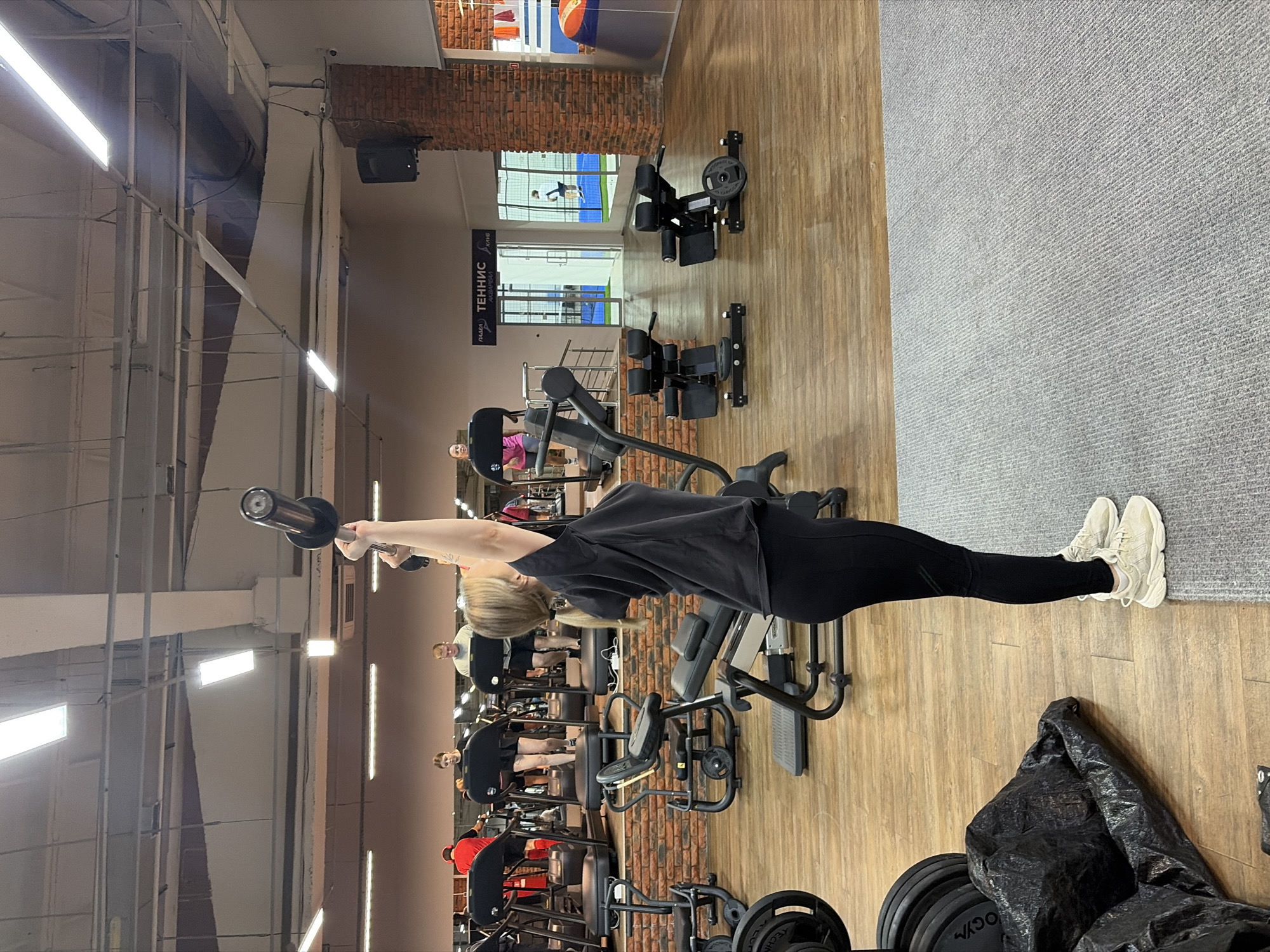 A woman in a black shirt and black leggings is standing and holding a barbell over her head in a gym. She is wearing white sneakers. There are weight machines in rows behind her. The floor is wood with a gray carpet in one section. There is a brick wall in the background, and a window overlooking an indoor tennis court. A sign in Russian says "Tennis." Based on the sign and the available information, the location is likely a gym in Chaikovsky, Russia that has a tennis court.